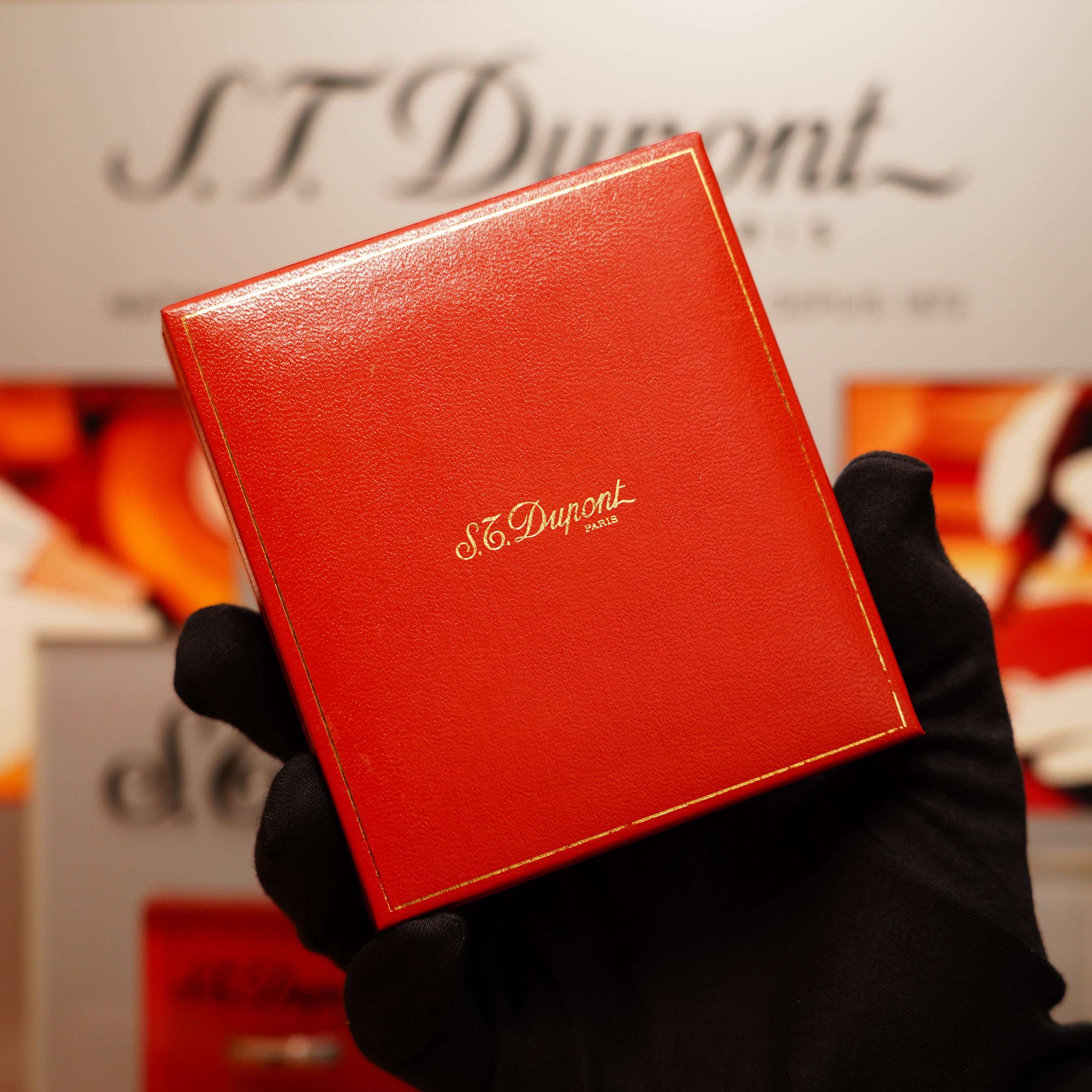 A gloved hand holding a red S.T. Dupont box with gold lettering for the Vintage 1990 St Dupont Overall Black Lacquer Early 24k Gold Ligne D Sealed Lighter.