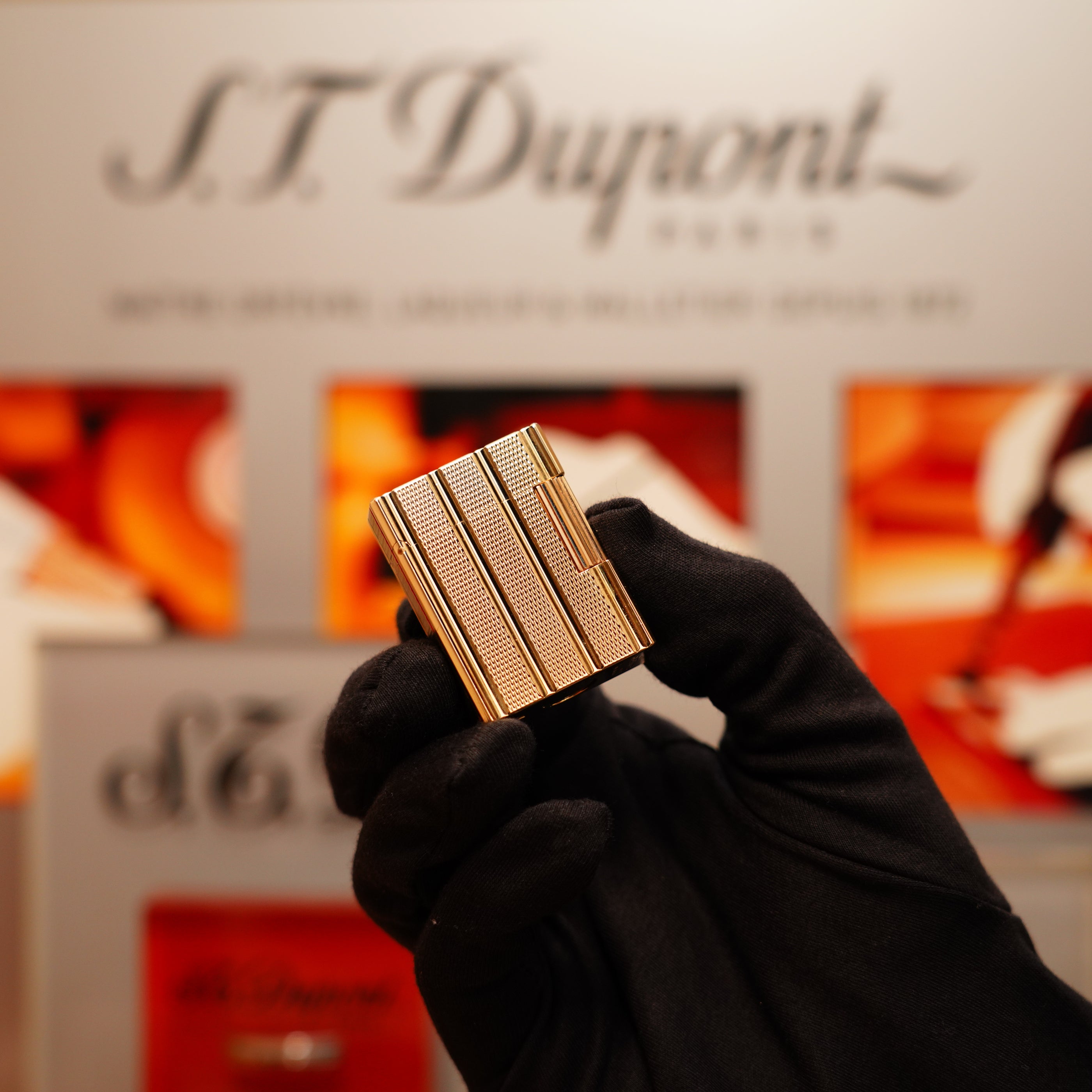 A gloved hand holds a Vintage 1990 S.T. Dupont 24k Gold Iconic Ligne 1 Lighter with a small dots and line pattern in front of a branded display.