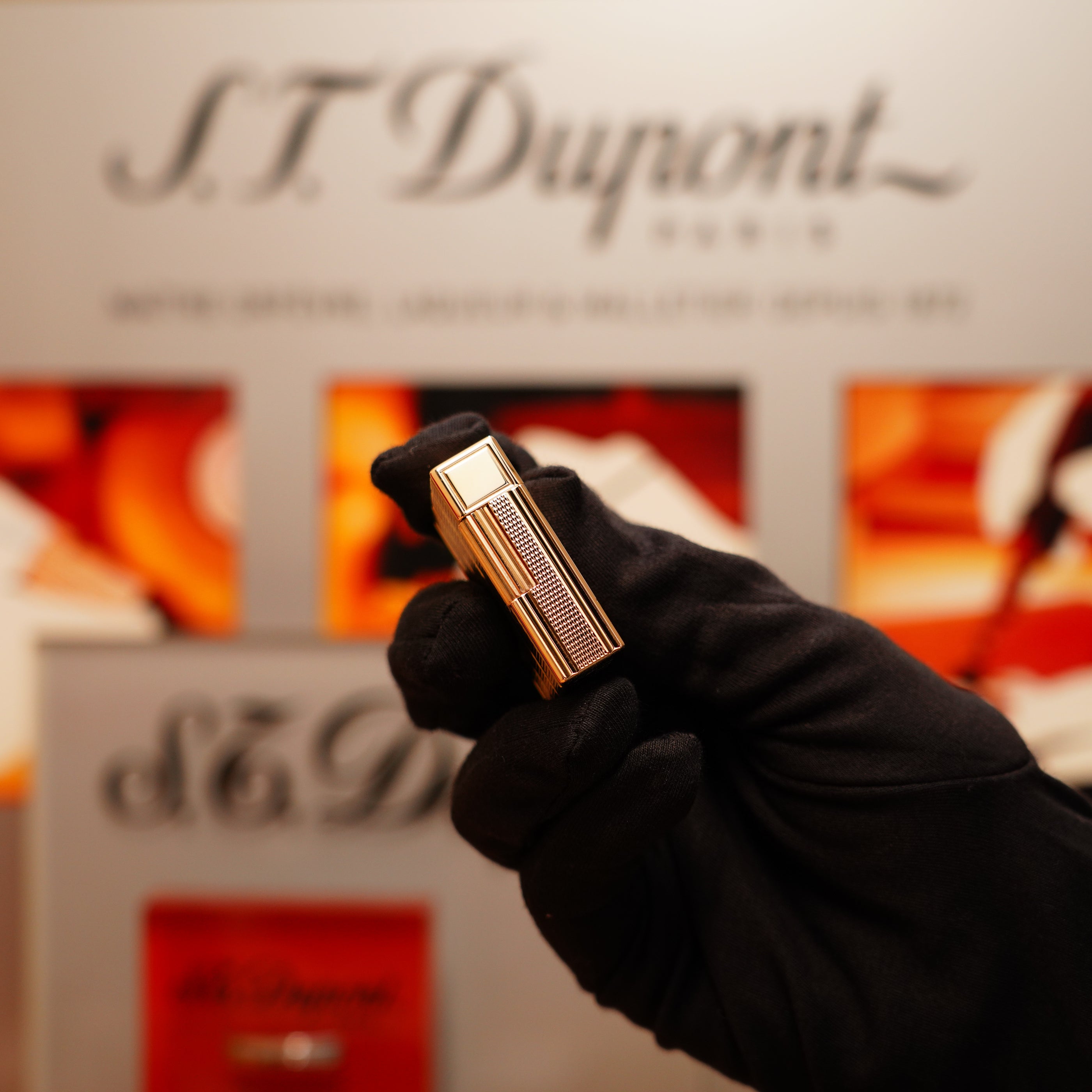 A gloved hand holding a Vintage 1990 S.T. Dupont 24k Gold Iconic Ligne 1 lighter with a small dots and line pattern in front of a blurred S.T. Dupont display.