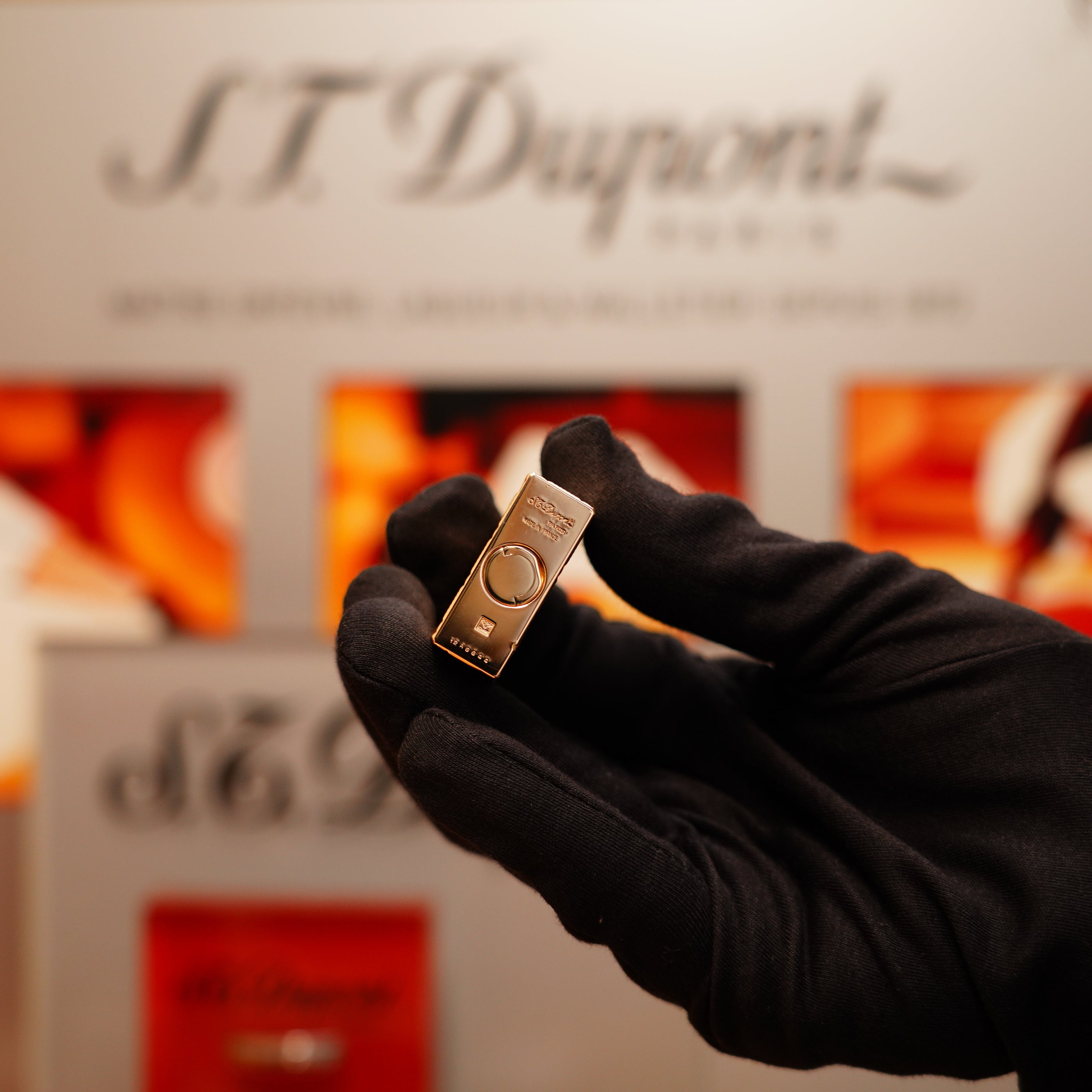 A gloved hand holds a small, rectangular, gold 24k St Dupont Ligne 1 lighter with a dots and line pattern in front of a blurred background adorned with "S.T. Dupont" branding.