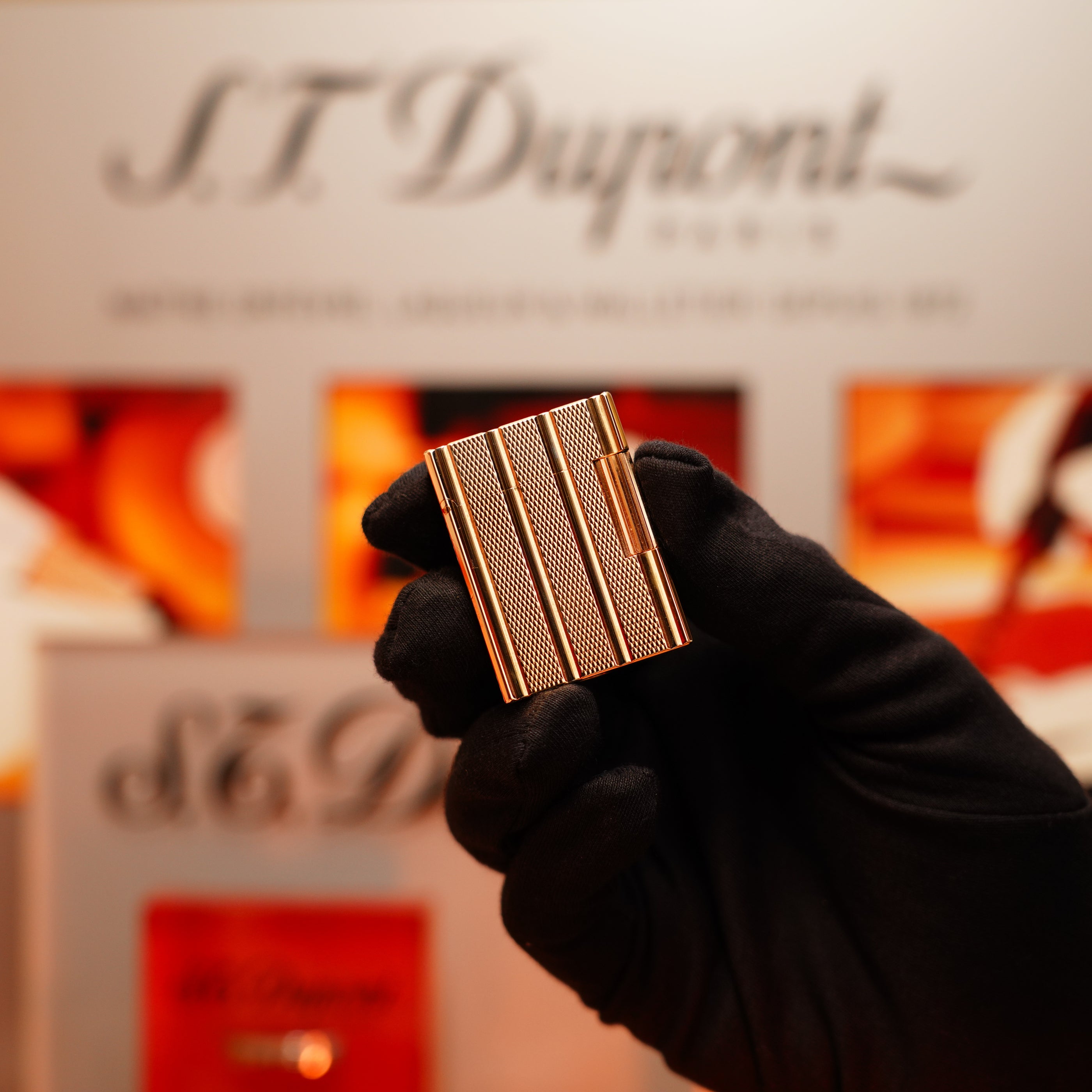 A gloved hand holds a Vintage 1990 S.T. Dupont 24k Gold Ligne 1 lighter with small dots and line pattern in front of a blurred S.T. Dupont display.
