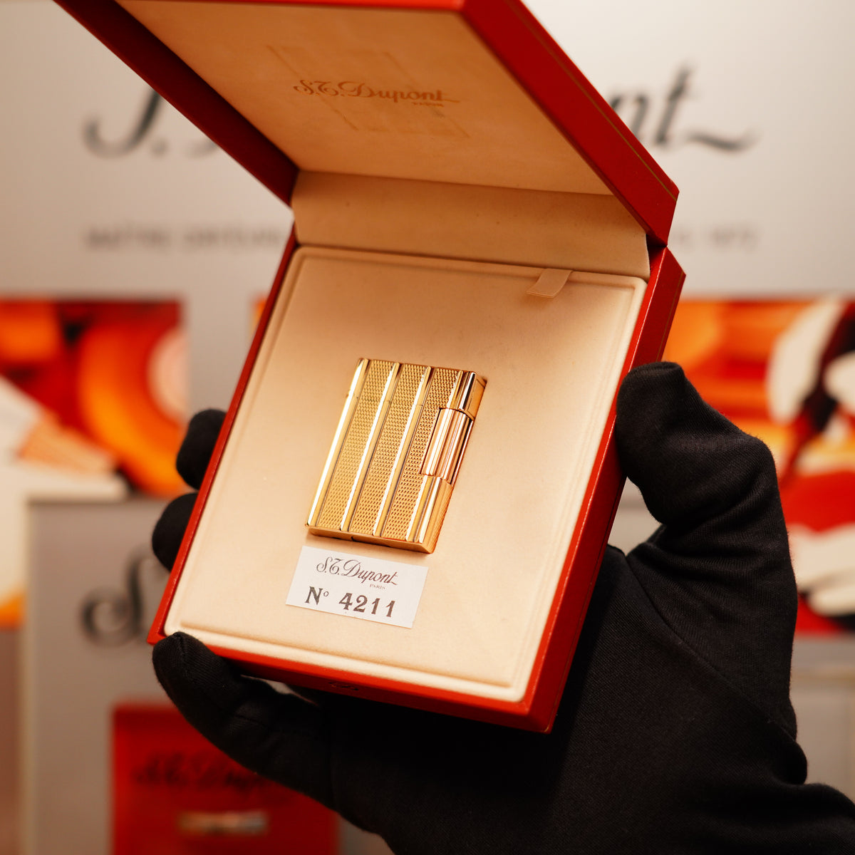 A gloved hand holds an open red box displaying a gold lighter with a black cap, labeled "Vintage 1990 St Dupont 24k Gold Iconic Ligne 1 Small Dots and Line Pattern Lighter," against a blurred background.