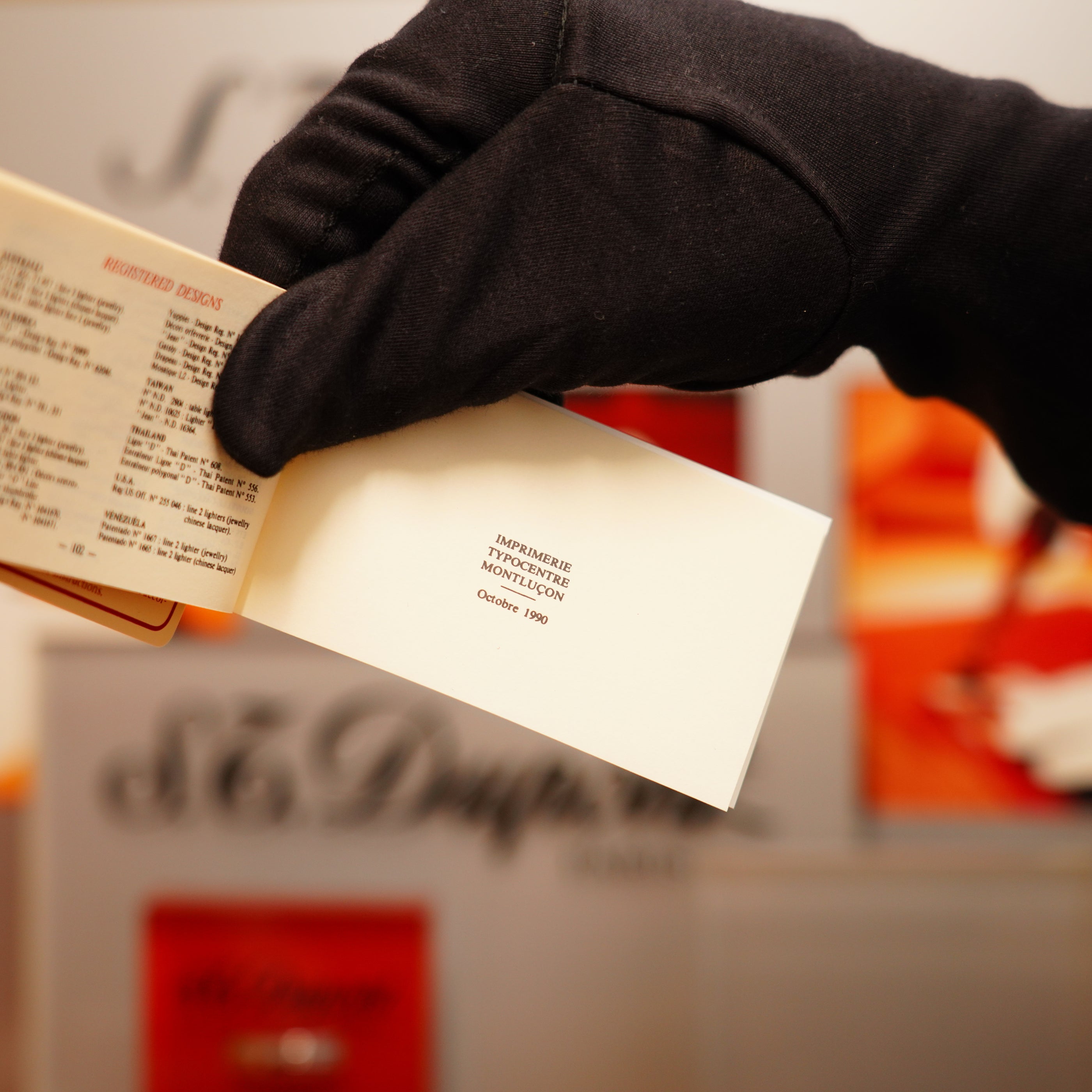 A gloved hand holds an open booklet showing a printed 1999 date, with a blurred background and an S.T. Dupont vintage 1990 St Dupont 24k Gold Iconic Ligne 1 Small Dots and Line Pattern Lighter.