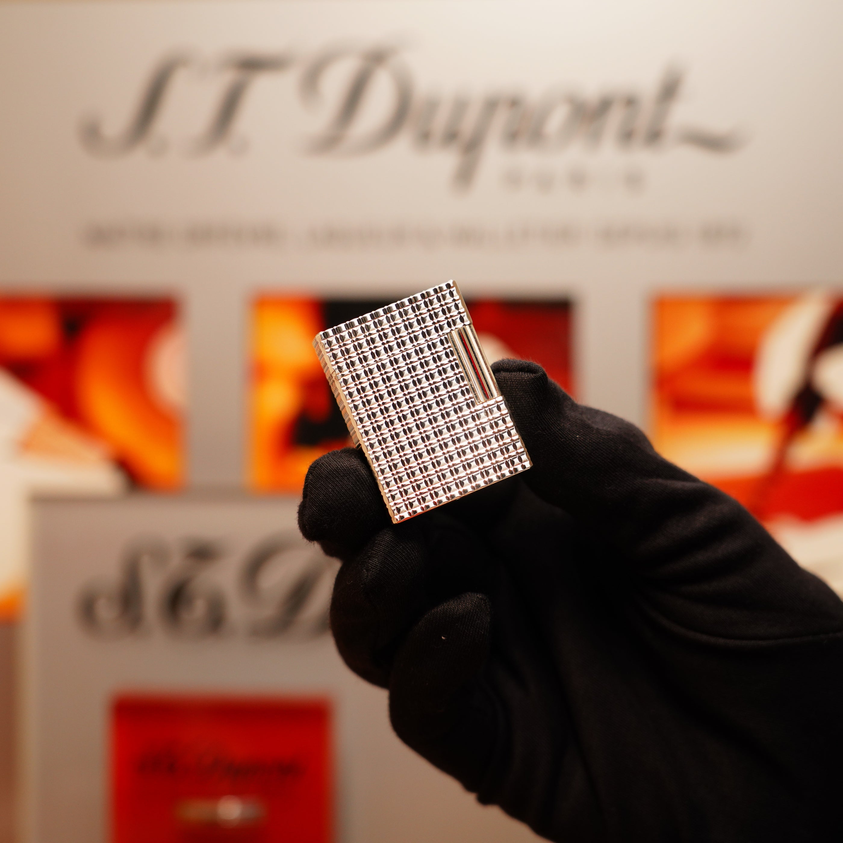 A gloved hand holds a silver lighter with a rare window pattern against a blurred background showing "S.T. Dupont.