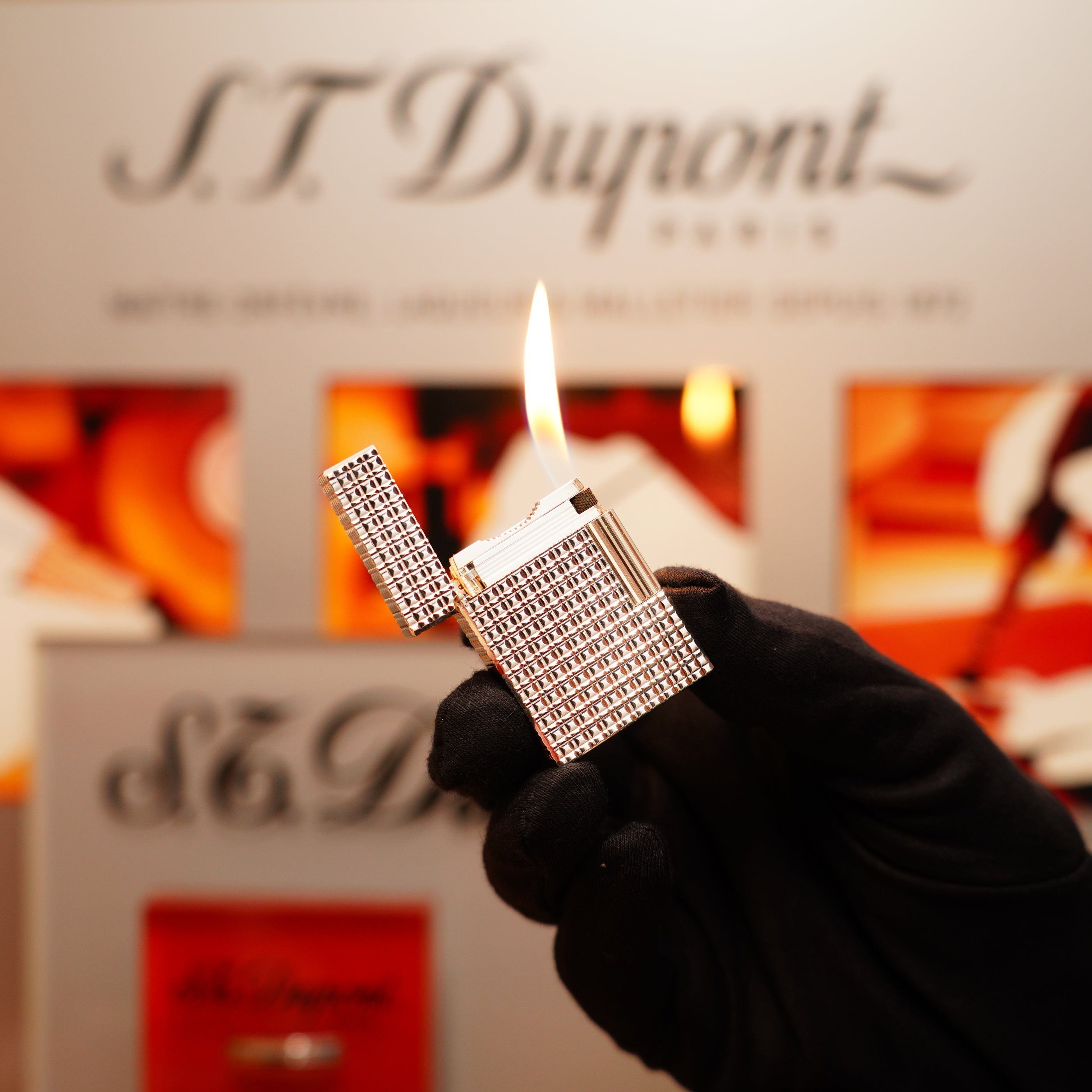 A gloved hand holds a lit Vintage 1990 St Dupont Heavy Silver Finish Iconic Ligne 1 Rare Window Pattern Lighter in front of a blurred background, with "S.T. Dupont" visible.