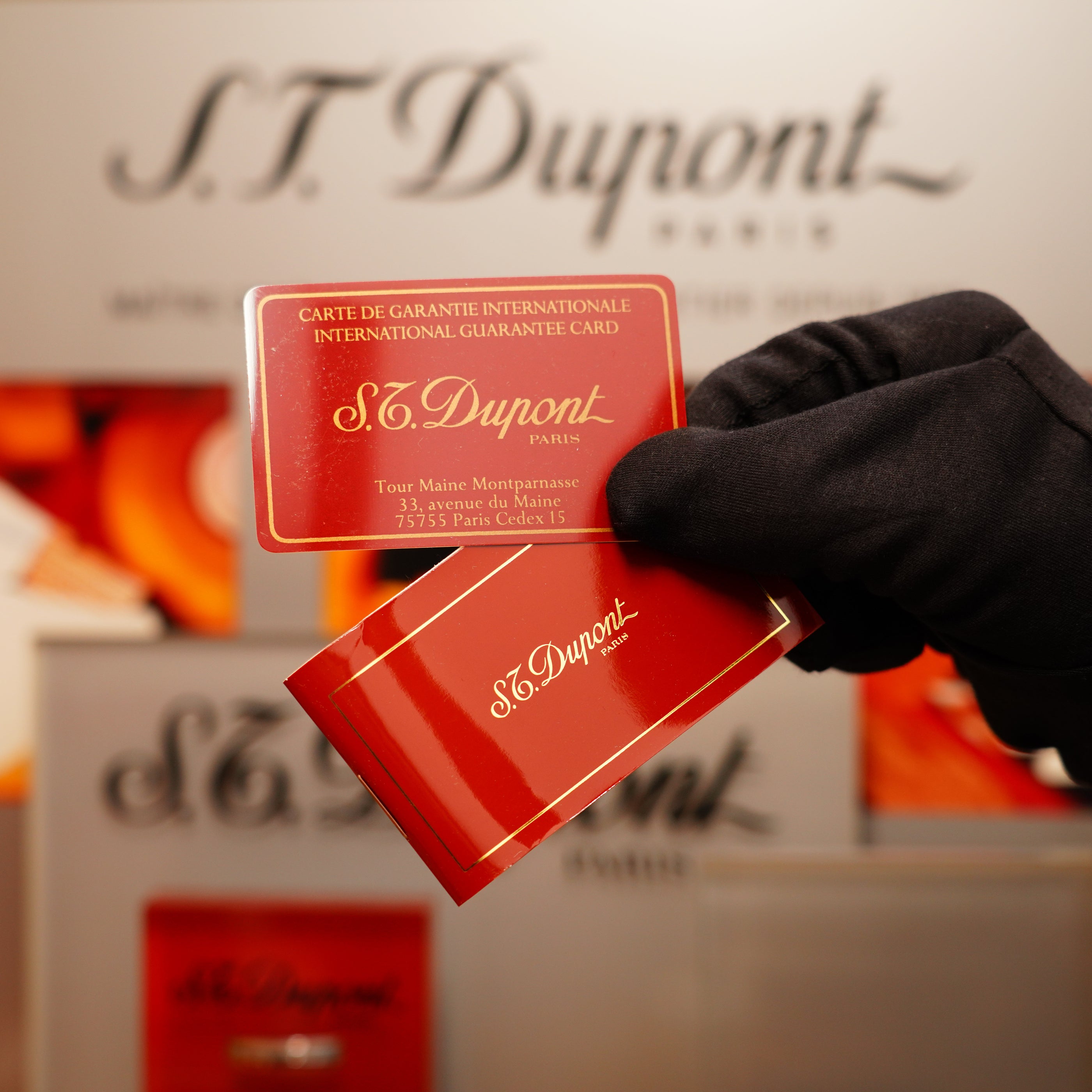 A gloved hand holds an S.T. Dupont international guarantee card and a matching red envelope, set against a blurred backdrop emphasizing the S.T. Dupont branding, highlighting the Vintage 1990 St Dupont Heavy Silver Finish Iconic Ligne 1 Rare Window Pattern Lighter.