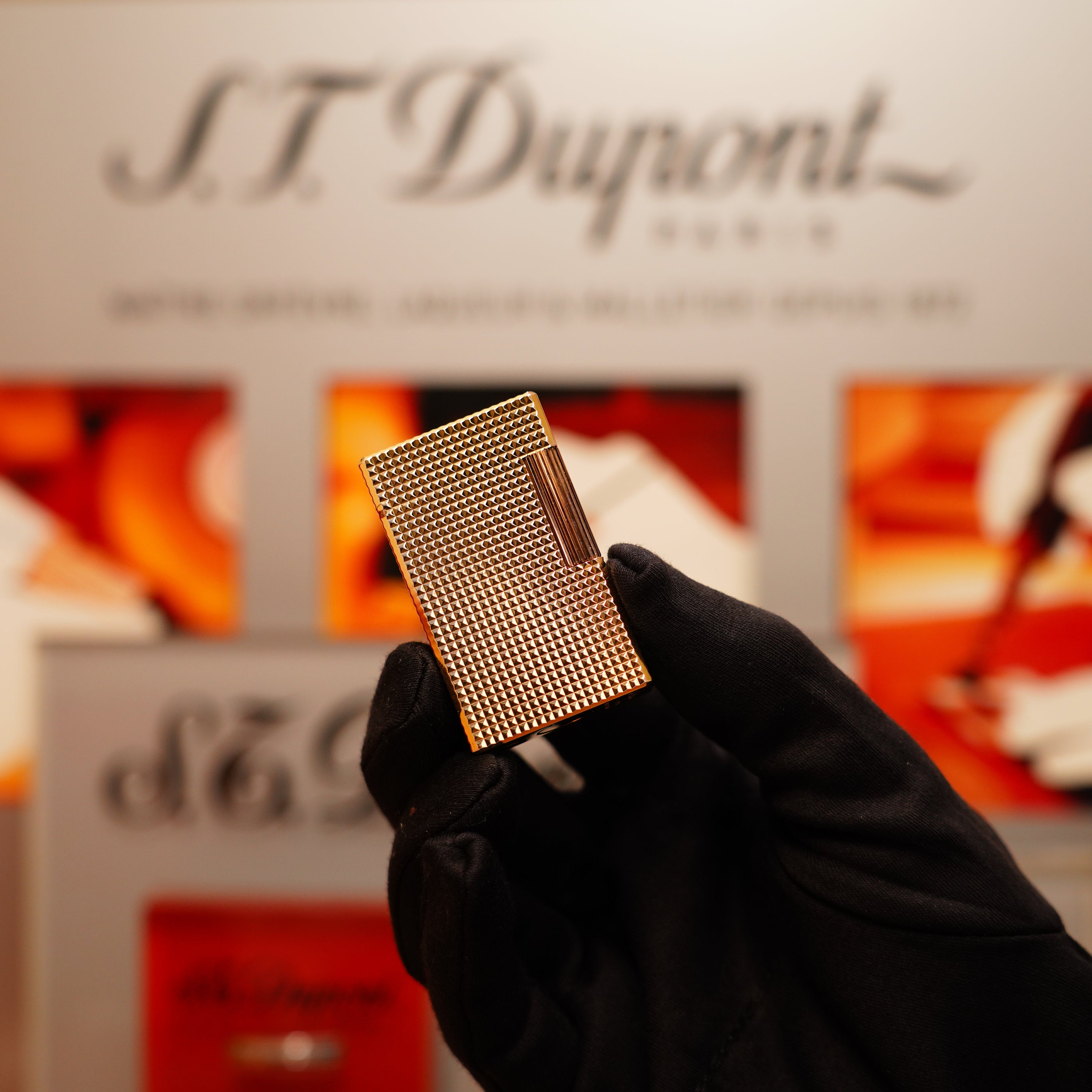 A gloved hand holds a Vintage 1990 S.T. Dupont 24k Gold Iconic Ligne 1 Diamond Head Pattern Lighter in front of a blurred S.T. Dupont sign.