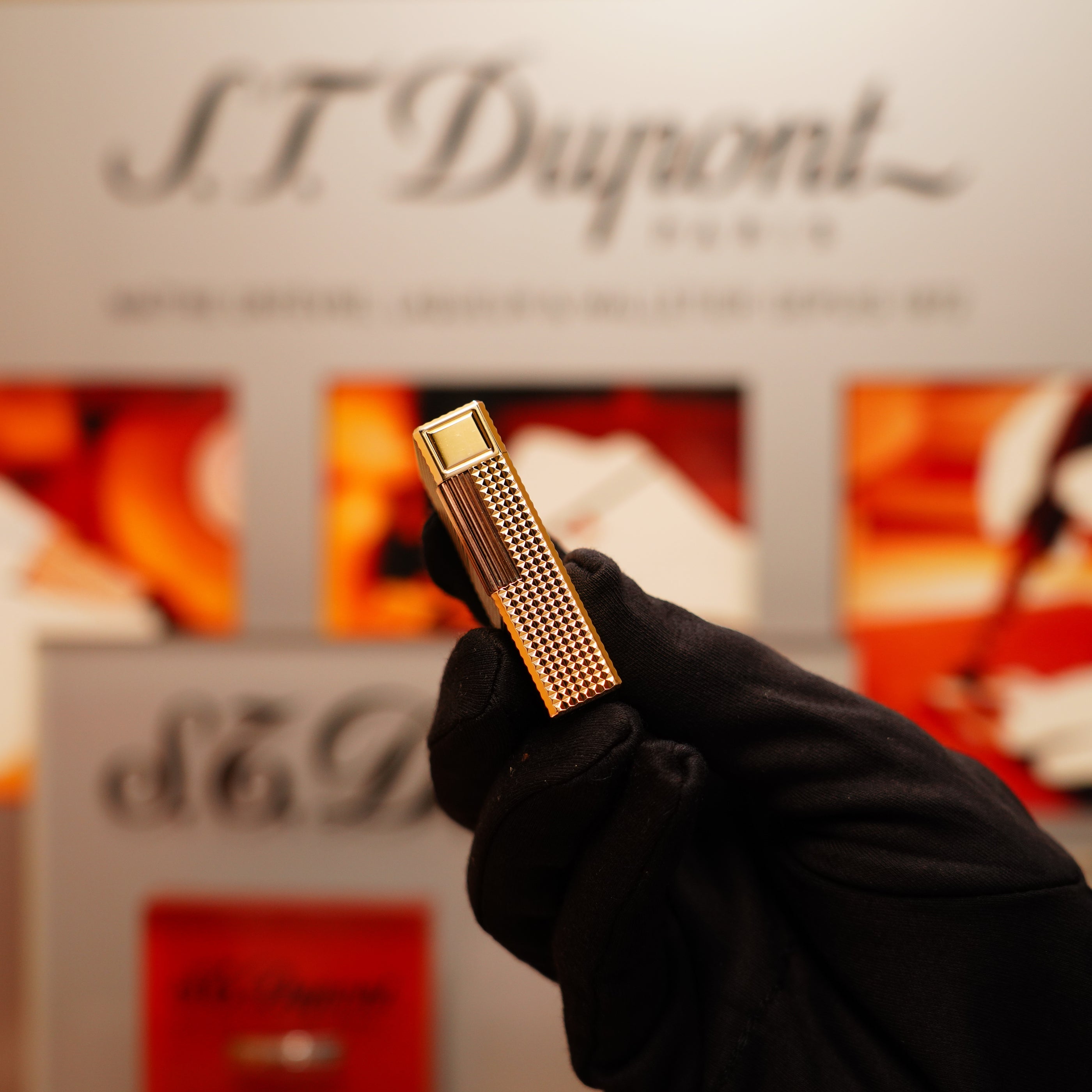 A gloved hand holds a Vintage 1990 S.T. Dupont 24k Gold Iconic Ligne 1 Diamond Head Pattern Lighter. Blurred background highlights the "S.T. Dupont" branding and imagery.