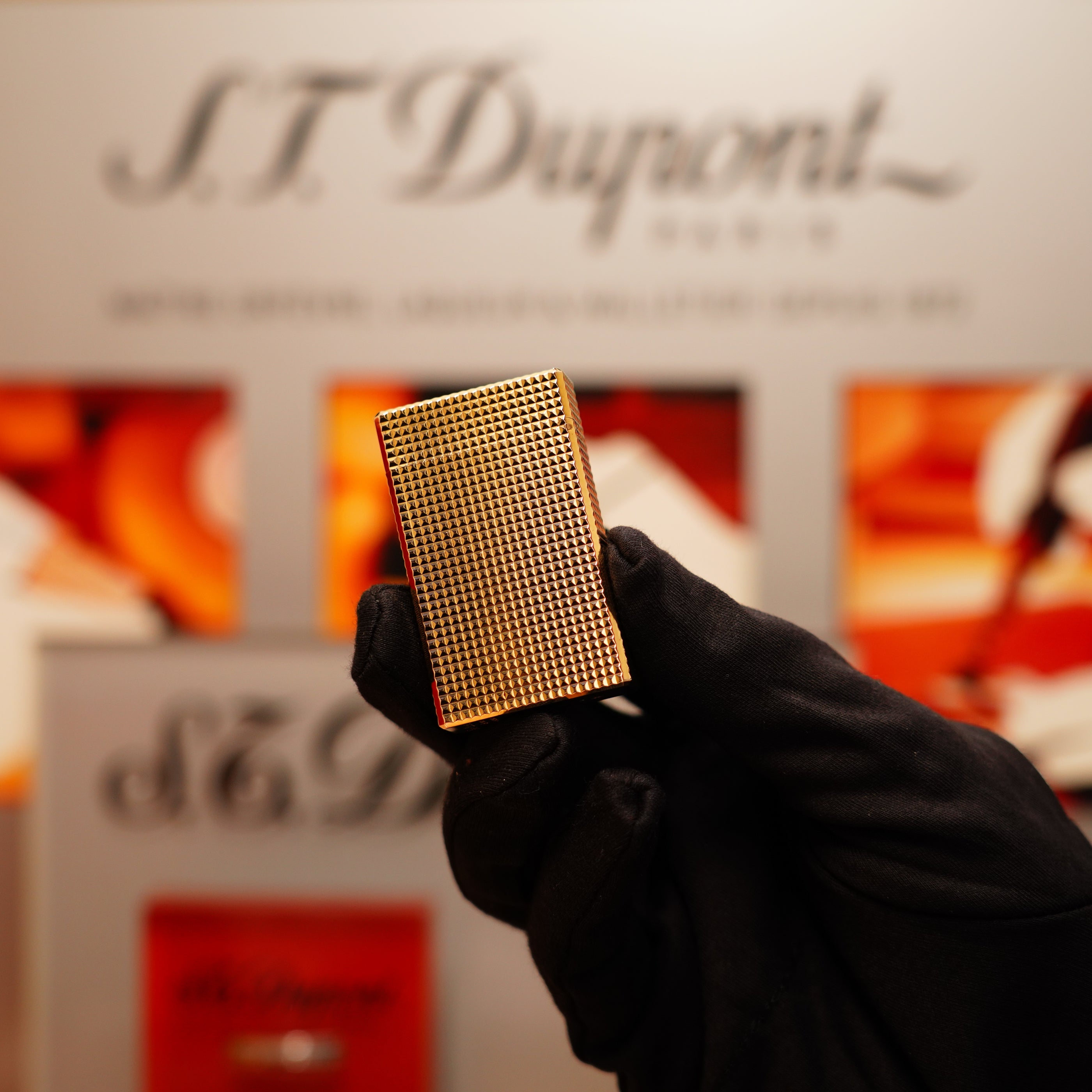 A gloved hand holds a Vintage 1990 S.T. Dupont 24k Gold Iconic Ligne 1 Diamond Head Pattern Lighter against a blurred background with soft text and warm colors.