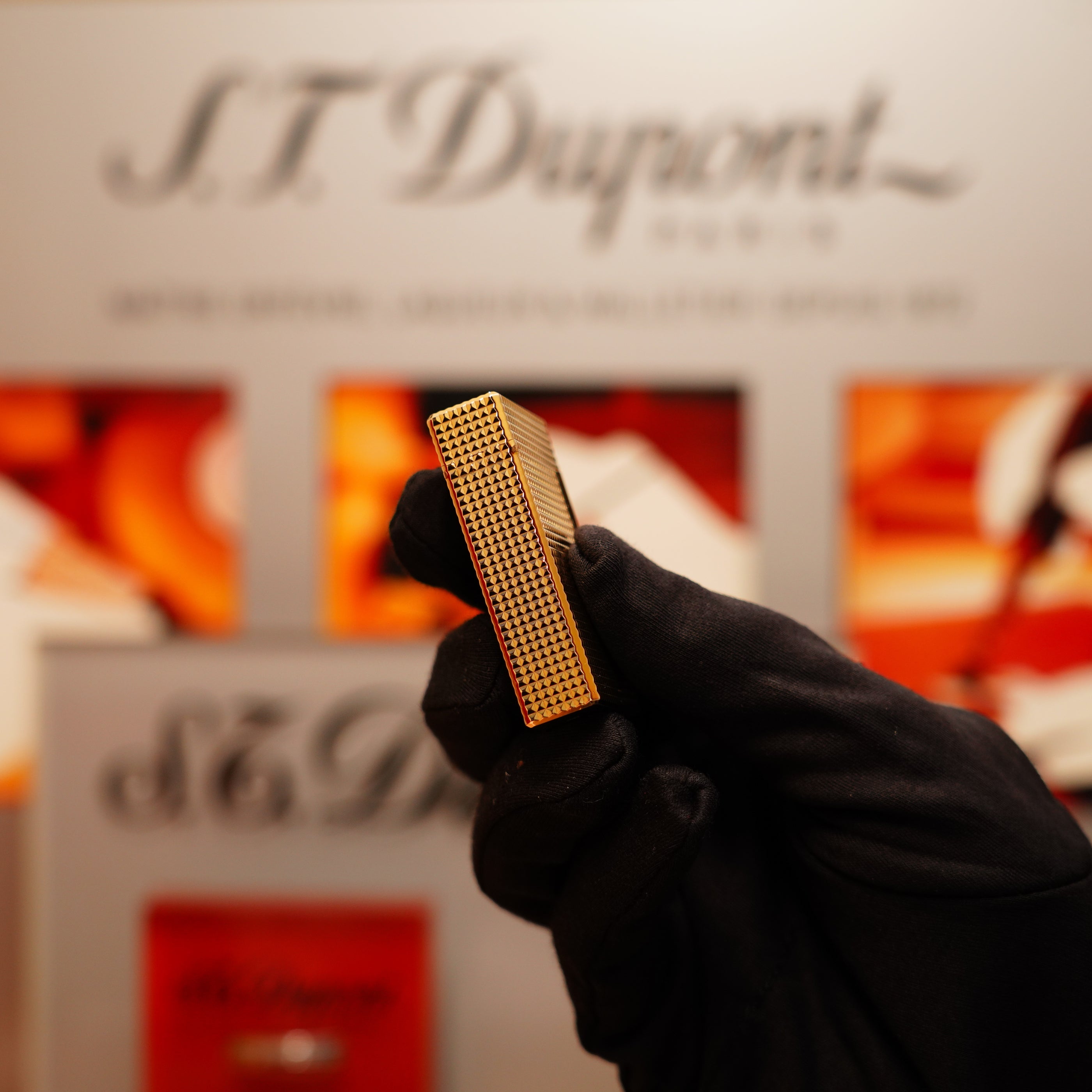 A gloved hand holds a detailed, rectangular Vintage 1990 S.T. Dupont 24k Gold Iconic Ligne 1 lighter with a Diamond Head pattern, against a blurred branded backdrop.