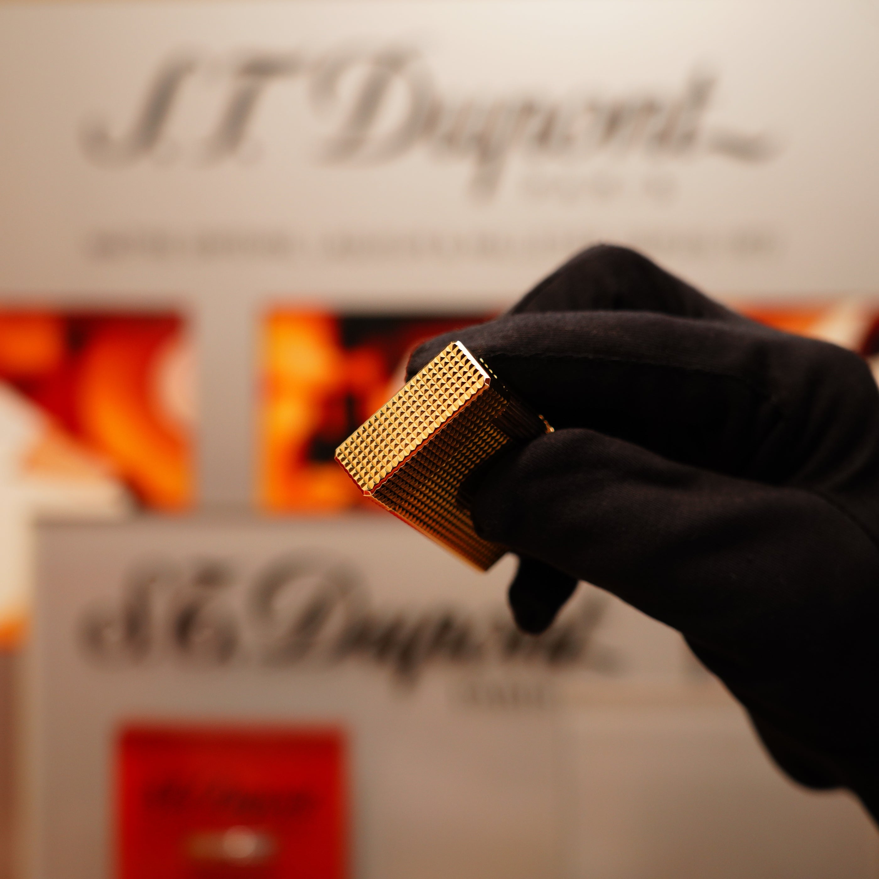 A gloved hand holds a Vintage 1990 S.T. Dupont 24k Gold Iconic Ligne 1 Diamond Head Pattern Lighter in front of a blurred S.T. Dupont display.