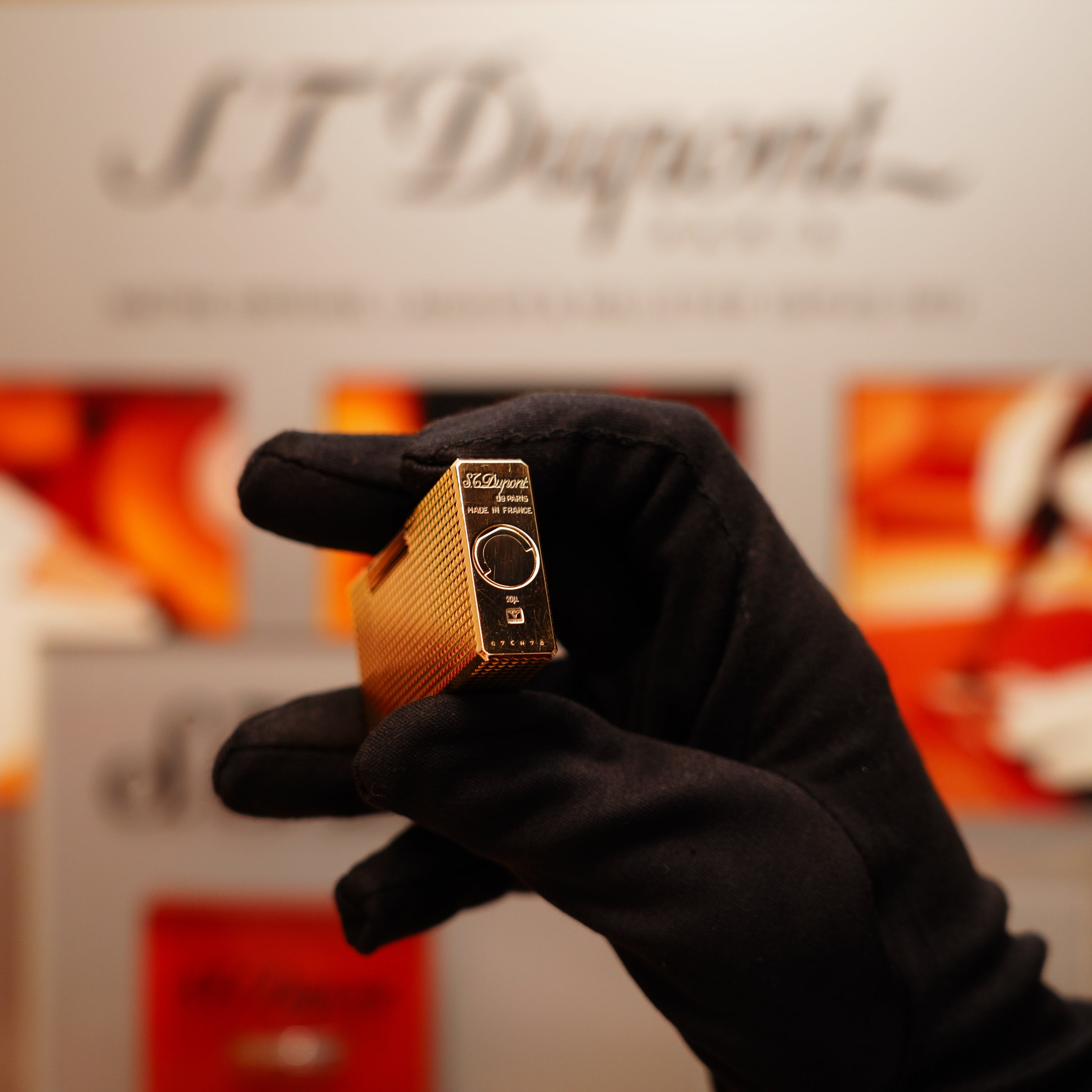 A gloved hand holds a Vintage 1990 S.T. Dupont 24k Gold Iconic Ligne 1 Diamond Head Pattern Lighter, with an S.T. Dupont branded background.