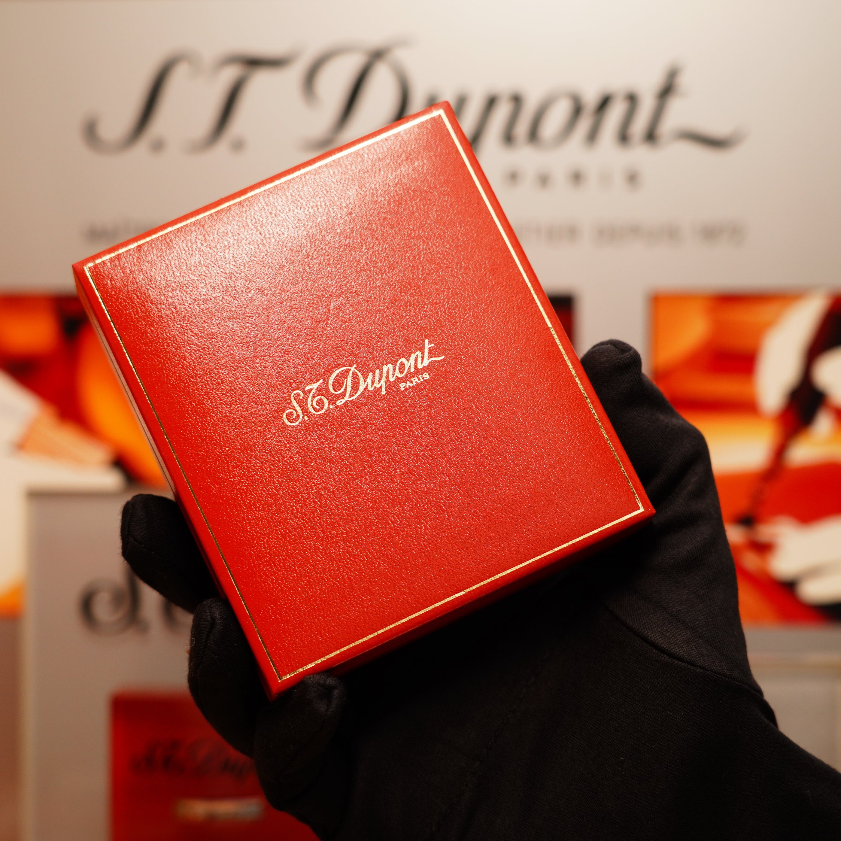 A gloved hand holds a red box with gold lettering reading "S.T. Dupont," containing a Vintage 1990 St Dupont 24k Gold Iconic Ligne 1 Diamond Head Pattern Lighter, set against a blurred background of similar branding.