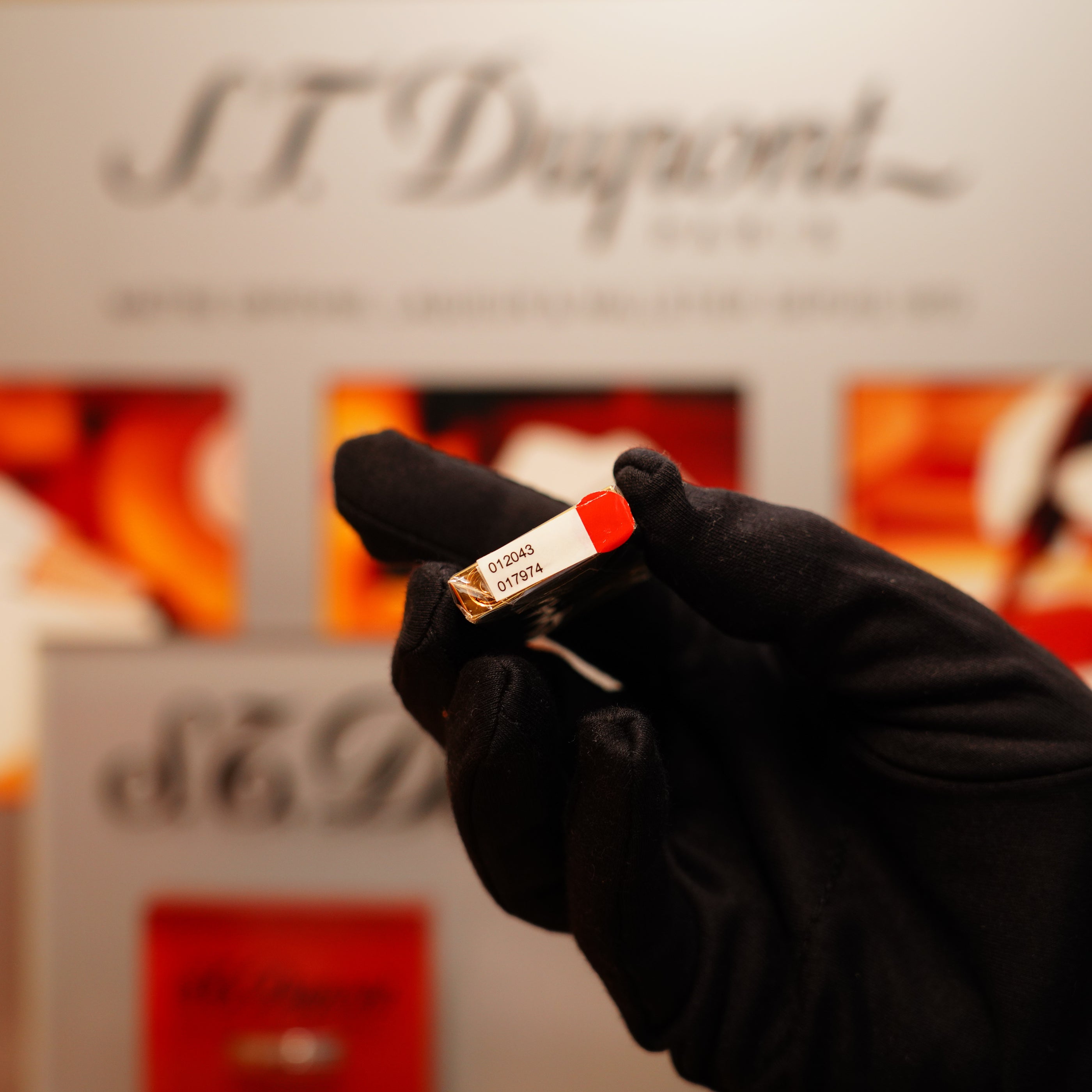 A gloved hand holds a small, red rectangular object with a label, against a blurred backdrop displaying "S.T. Dupont," likely showcasing the Vintage 1990 St Dupont Overall Black Lacquer Early 24k Gold Ligne D Sealed Lighter.