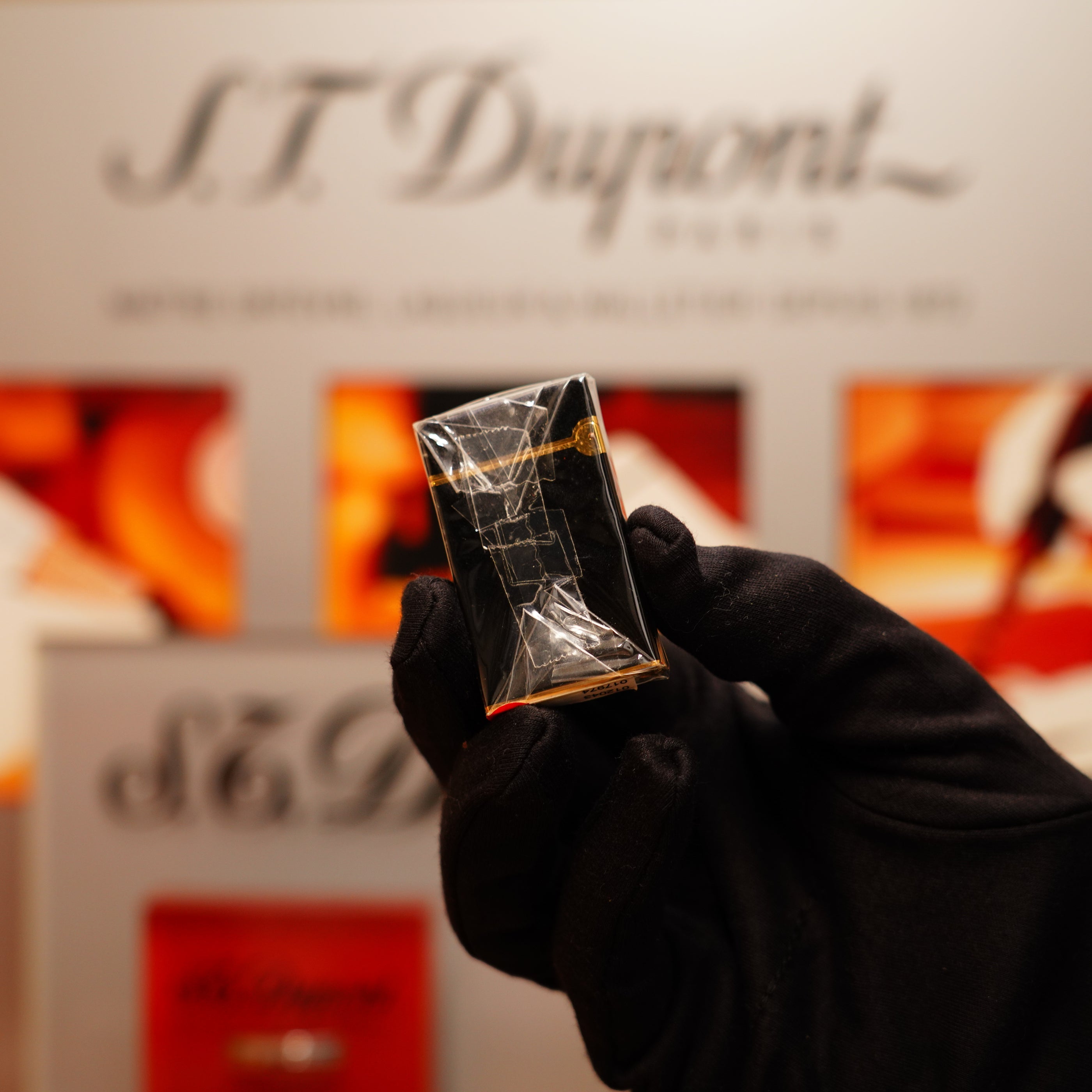 A gloved hand holds a vintage 1990 S.T. Dupont overall black lacquer and early 24k gold Ligne D lighter against a blurred "S.T. Dupont" branded background.