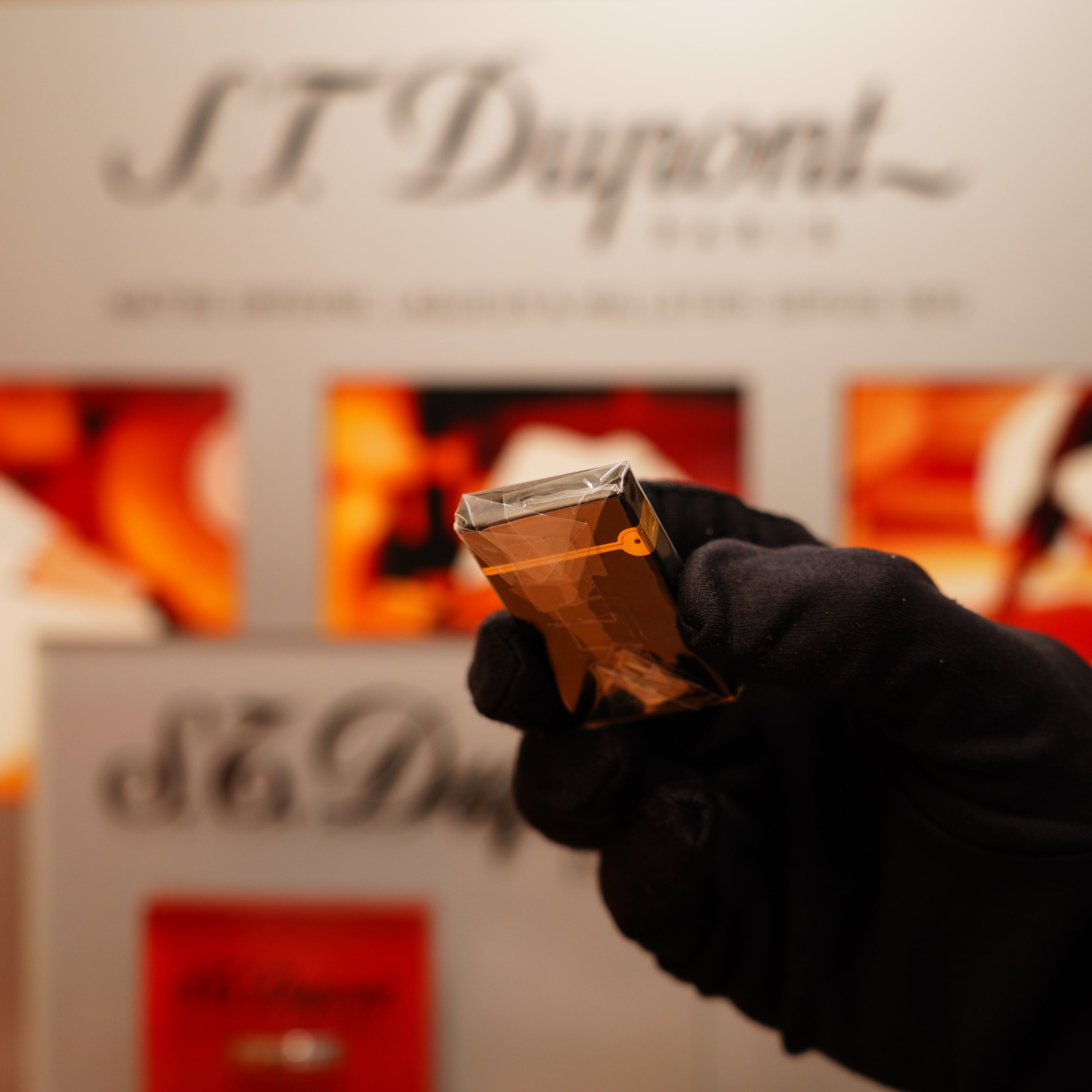 A gloved hand holds a small rectangular amber object with the S.T. Dupont display blurred in the background; it's the Vintage 1990 S.T. Dupont Overall Black Lacquer Early 24k Gold Ligne D Sealed Lighter.