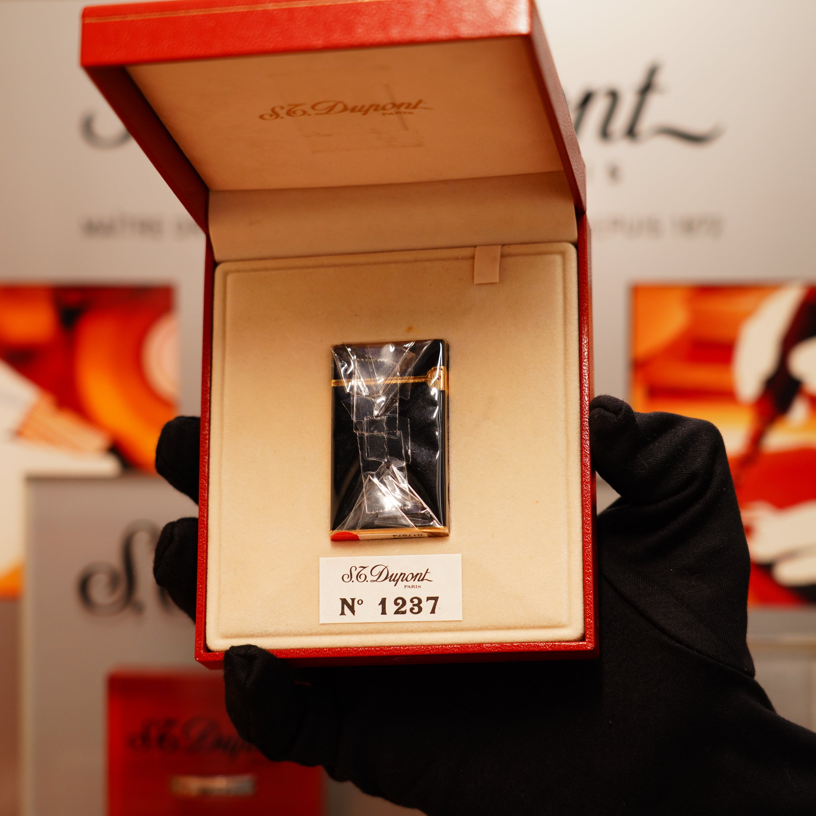 A gloved hand holds an open red box displaying a Vintage 1990 St Dupont Overall Black Lacquer Early 24k Gold Ligne D Sealed Lighter, "No 1287," from S.T. Dupont.