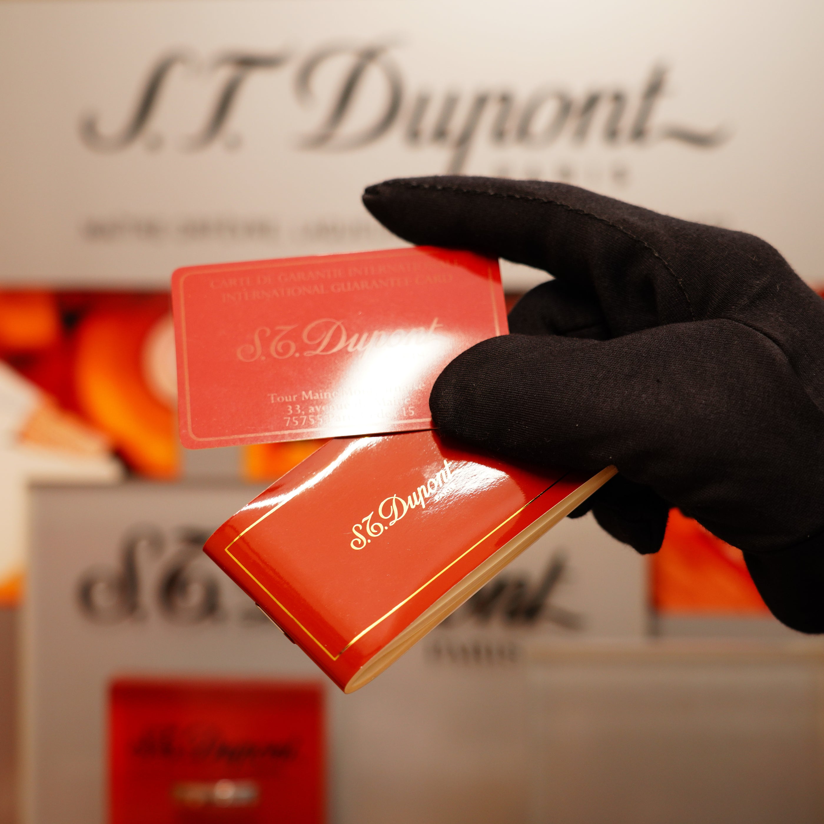 A gloved hand holds a Vintage 1990 S.T. Dupont Overall Black Lacquer Early 24k Gold Ligne D Sealed Lighter and card in front of a branded display.
