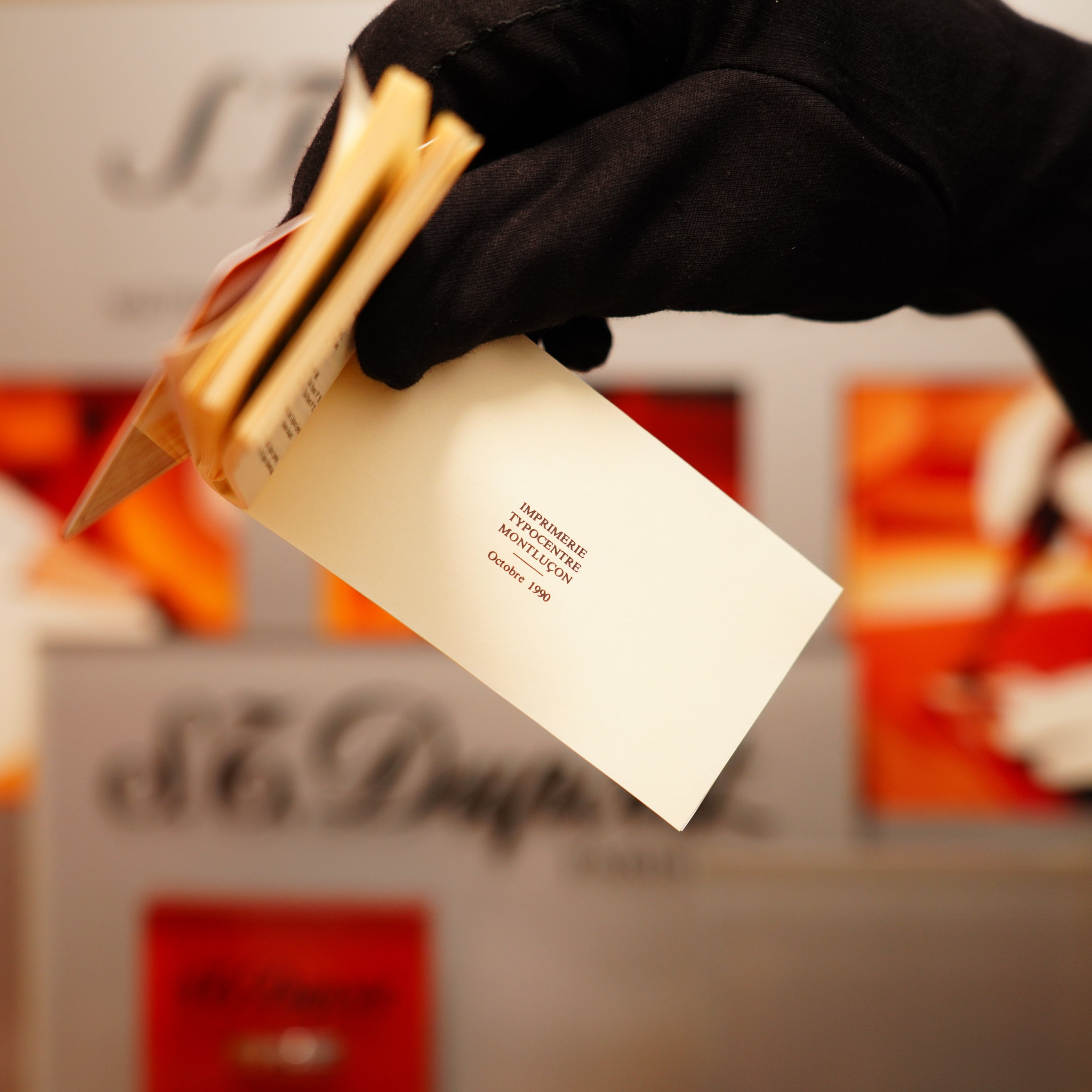 A gloved hand holds a small card reading "INTRAMUROS ARCHITECTURE October 1997" against a blurred backdrop with hints of the S.T. Dupont Vintage 1990 Black Lacquer and Early 24k Gold Ligne D Sealed Lighter visible.
