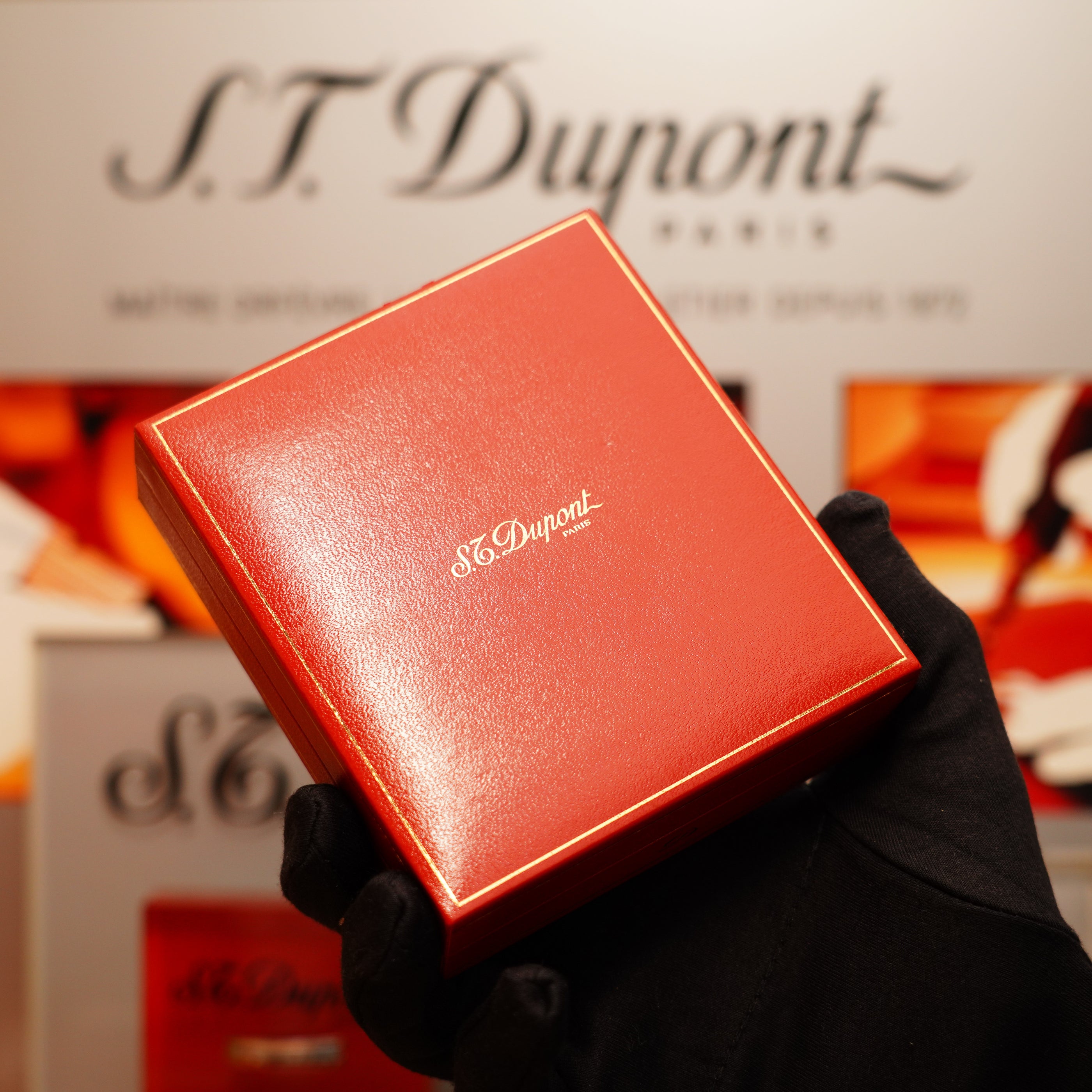 A gloved hand holds a red box with gold lettering containing a Vintage 1990 S.T. Dupont Overall Black Lacquer Early 24k Gold Ligne D Sealed Lighter, against a blurred background displaying the S.T. Dupont logo.