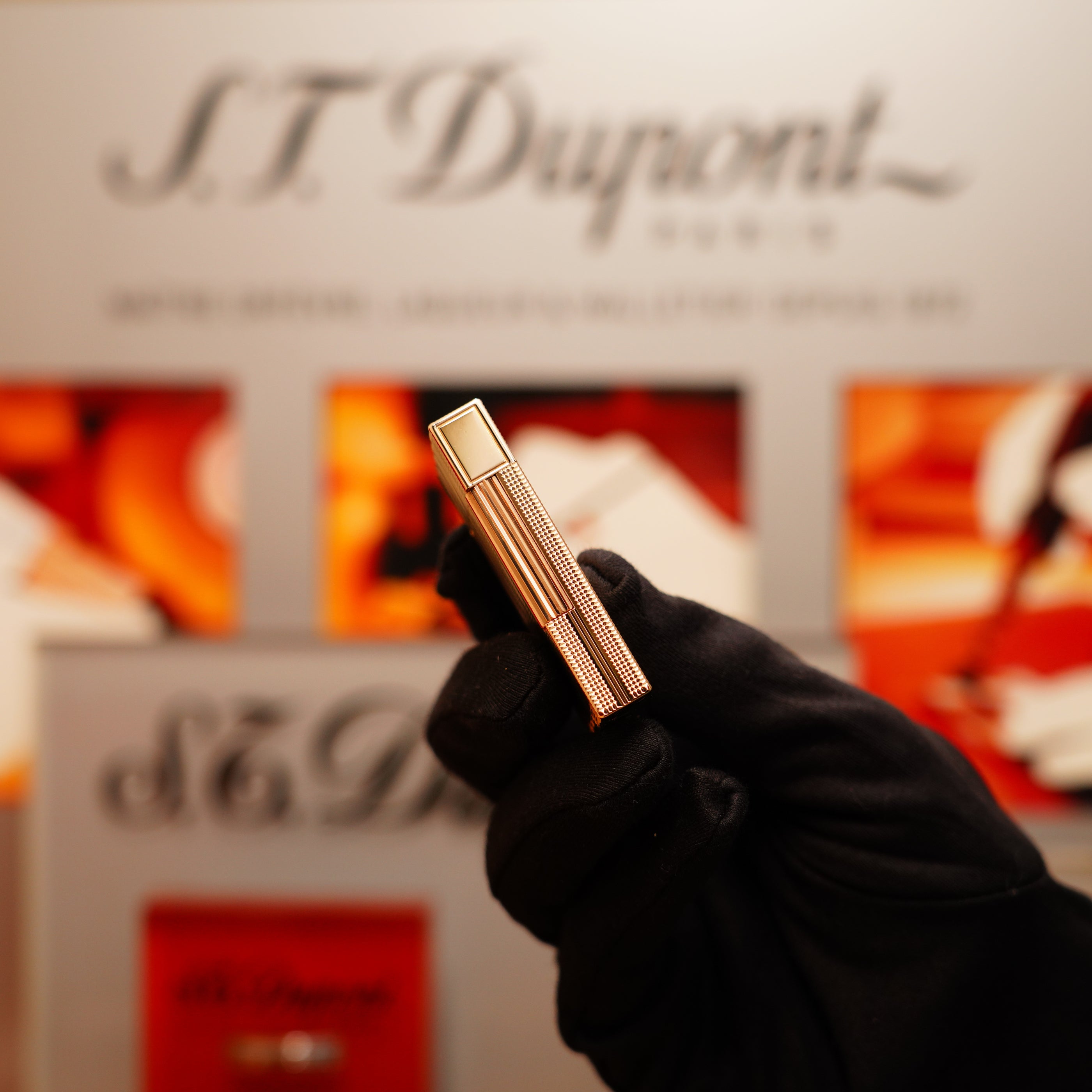 A gloved hand holds a Vintage 1978 S.T. Dupont 24k Gold Lignes Verticales Pointes Diamants Large Ligne 2 lighter with a metallic finish, against a blurred S.T. Dupont branded background.