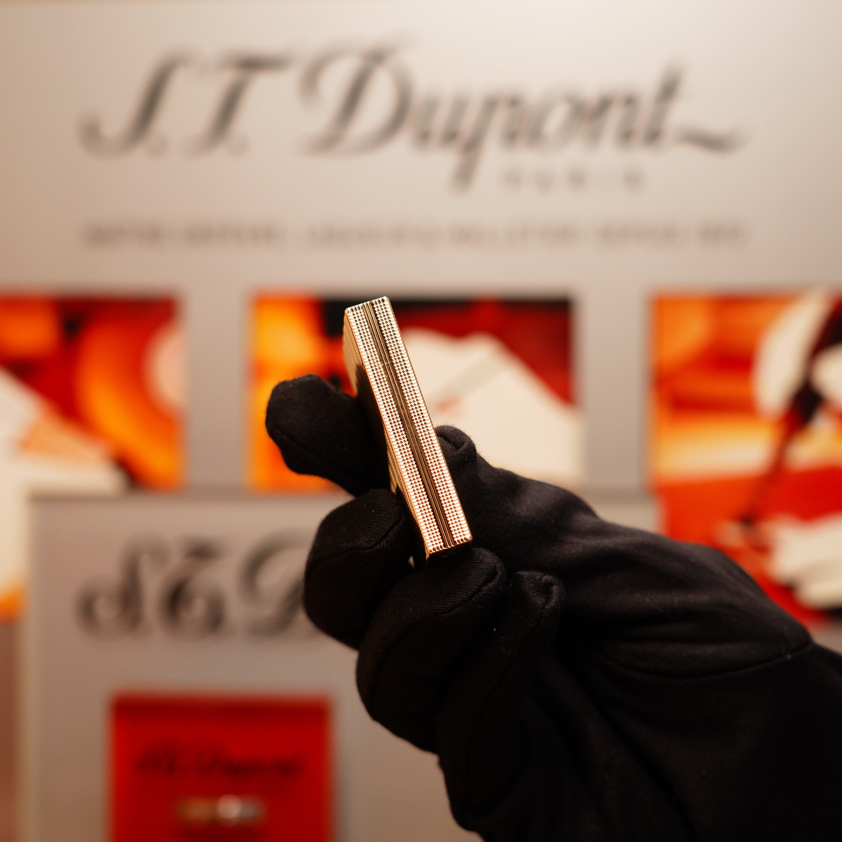 A gloved hand holds a Vintage 1978 S.T. Dupont Ligne 2 lighter in 24k gold with a ribbed design against a blurred backdrop featuring the brand name.