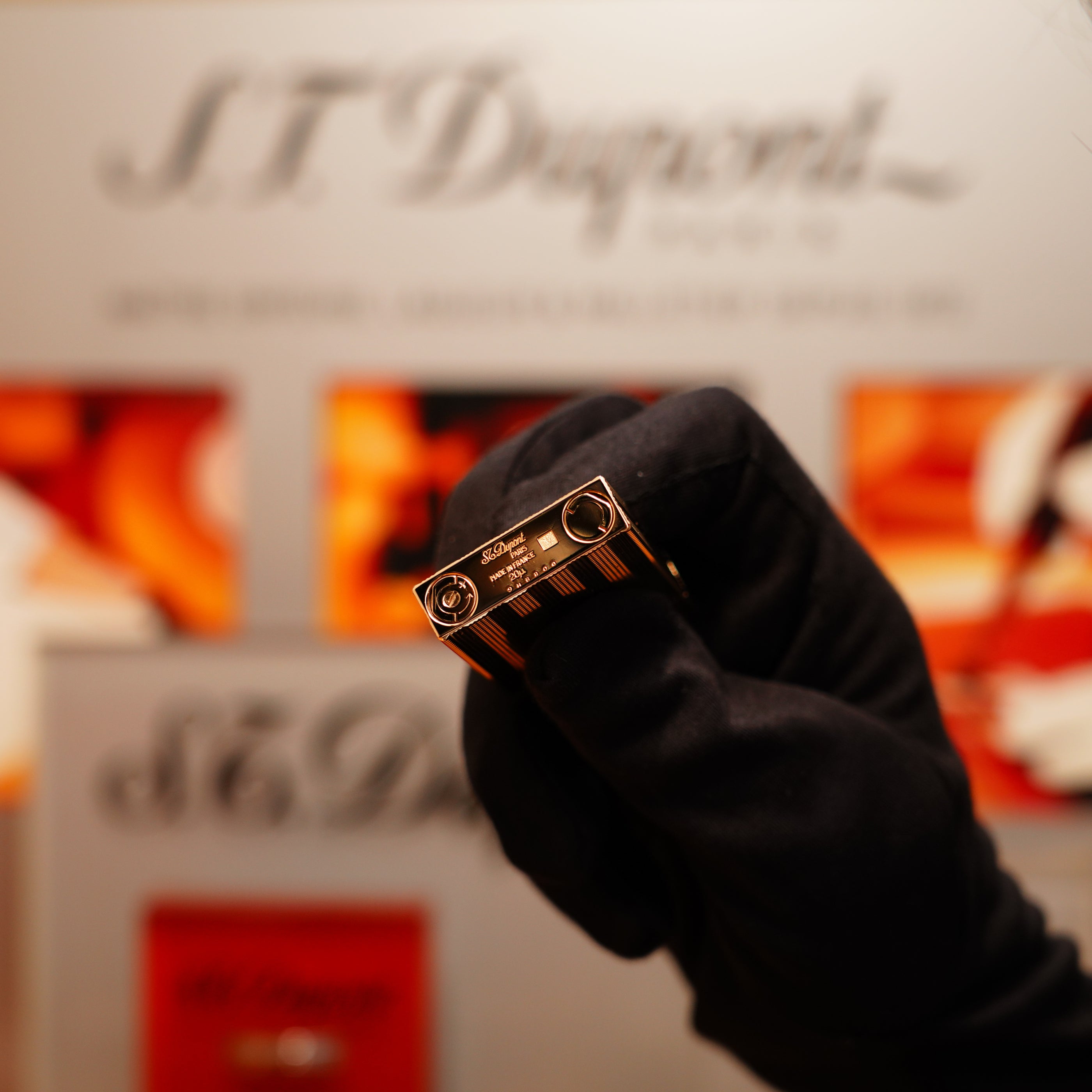 A gloved hand holds a Vintage 1978 S.T. Dupont 24k Gold Lignes Verticales Pointes Diamants Large Ligne 2 Lighter before a blurred display featuring the S.T. Dupont logo and luxury goods.