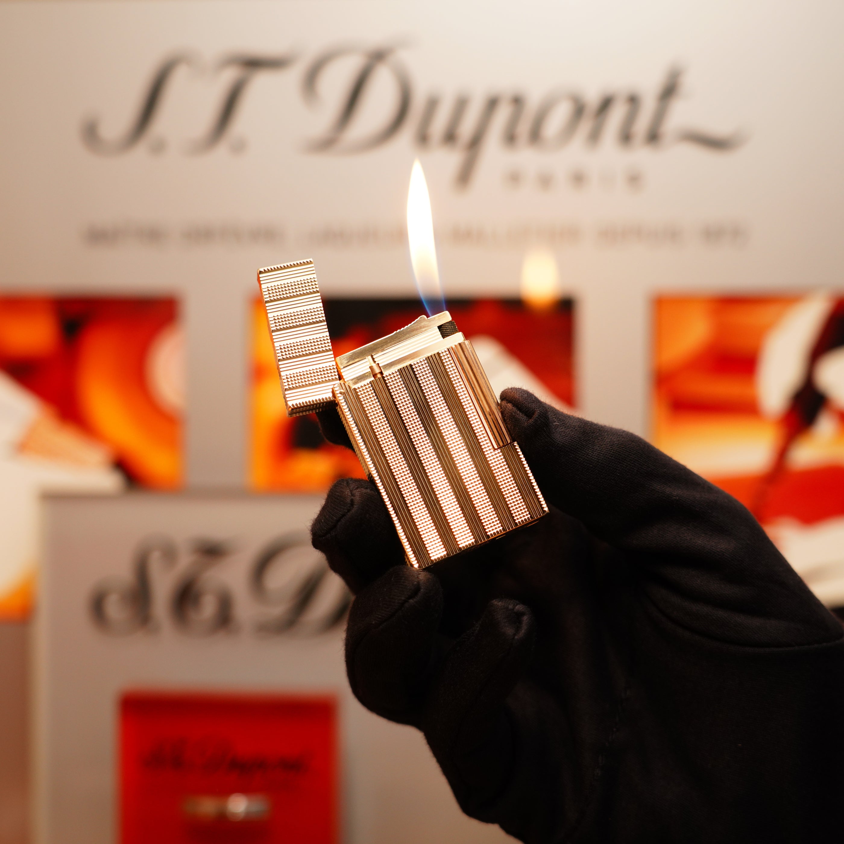 A gloved hand holds a lit Vintage 1978 S.T. Dupont 24k Gold Lignes Verticales Pointes Diamants Large Ligne 2 lighter, with a blurred background featuring the S.T. Dupont logo.
