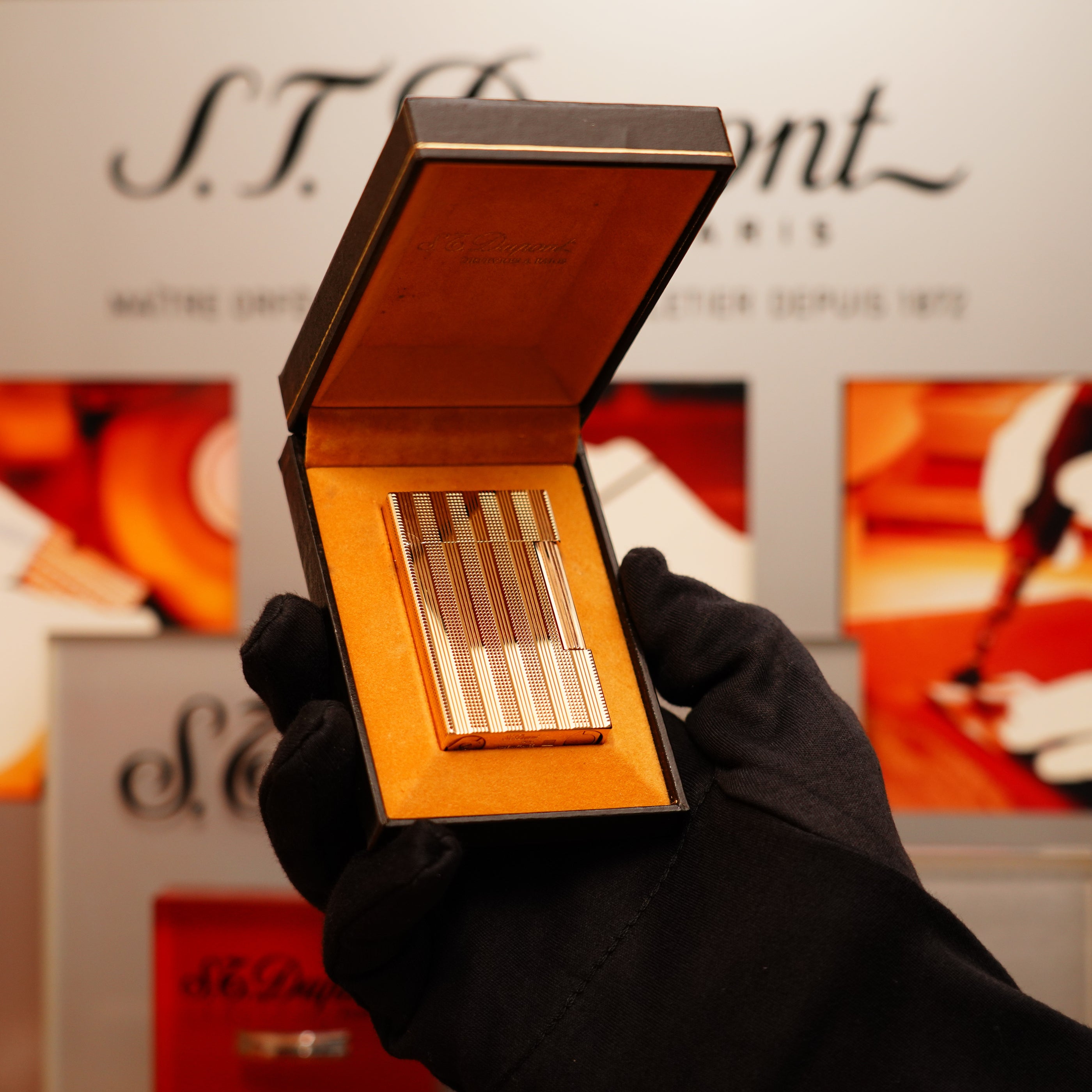 A gloved hand holds an open box containing a Vintage 1978 S.T. Dupont 24k Gold Lignes Verticales Pointes Diamants Lighter, with the S.T. Dupont brand name in the background.