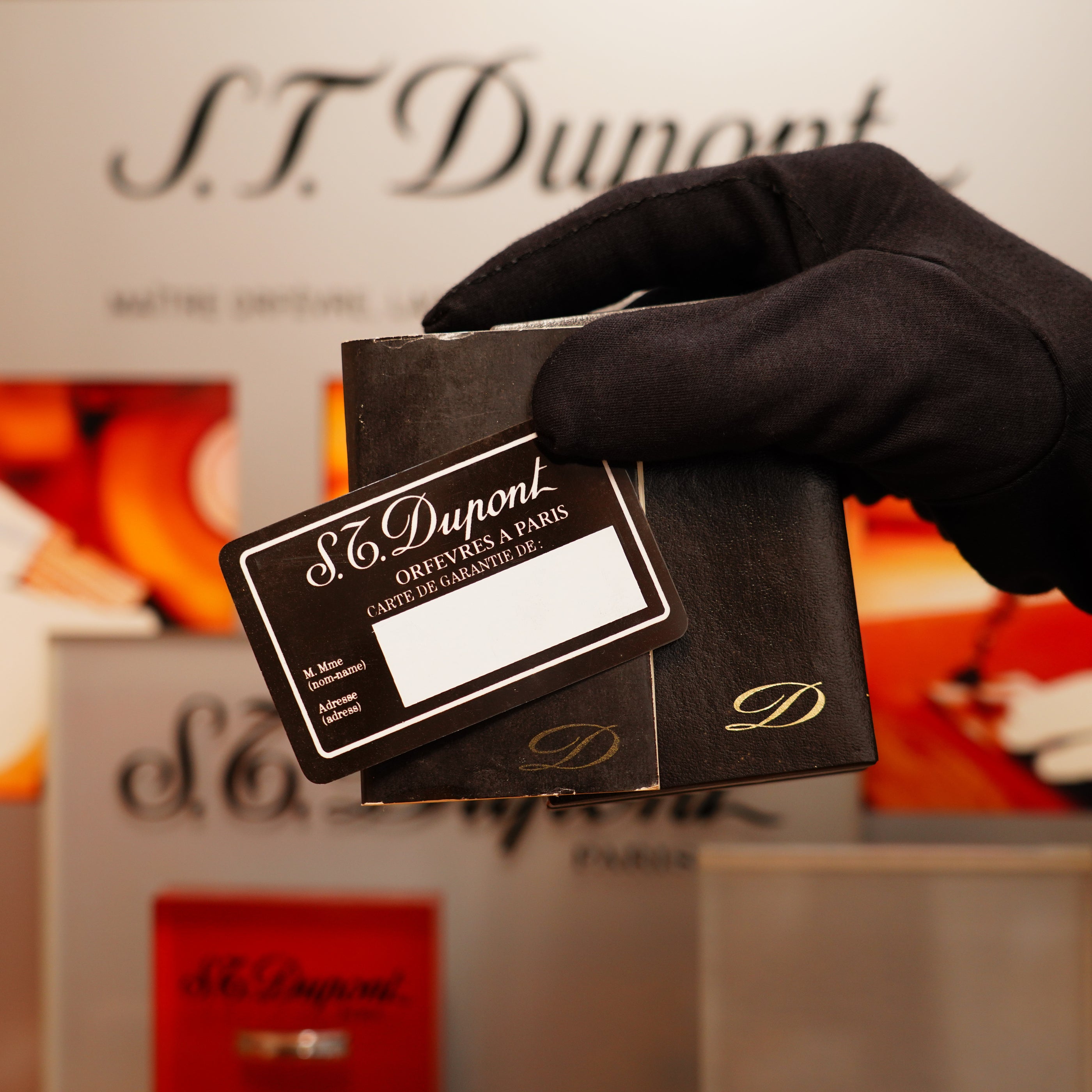 A gloved hand holds a black box containing a Vintage 1978 St Dupont 24k Gold Lignes Verticales Pointes Diamants Large Ligne 2 Lighter, with a card showing contact details. The blurred background features S.T. Dupont branding.