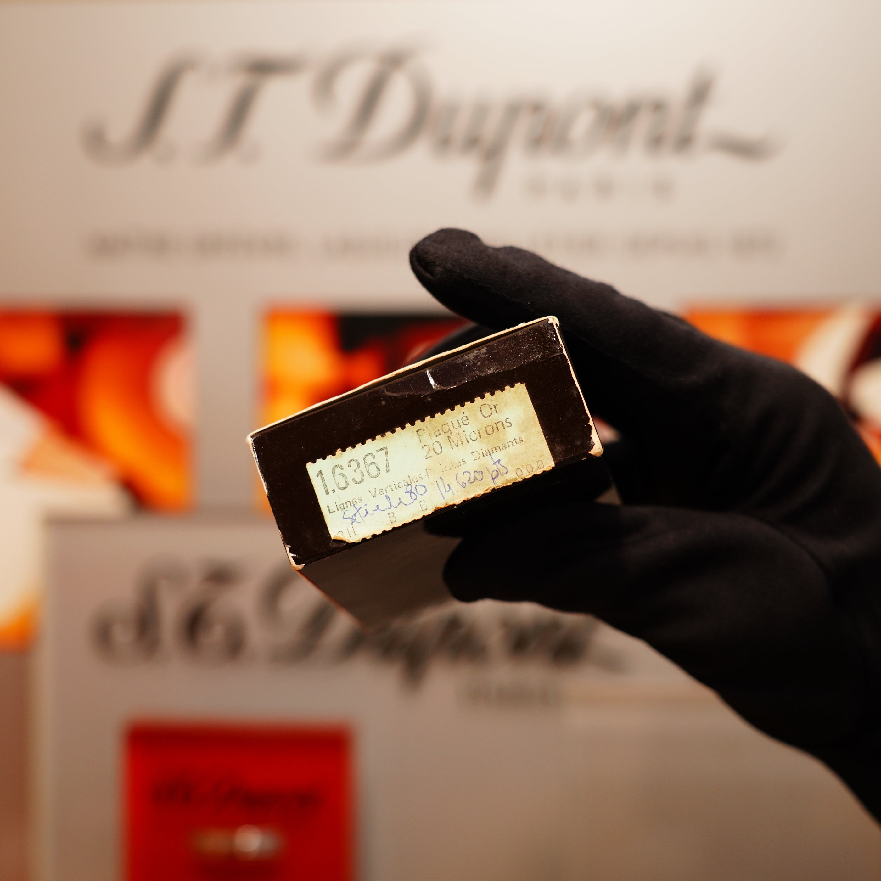 A gloved hand holds a small black box labeled "Vintage 1978 St Dupont 24k Gold Lignes Verticales Pointes Diamants Large Ligne 2 Lighter," set against a blurred S.T. Dupont background.
