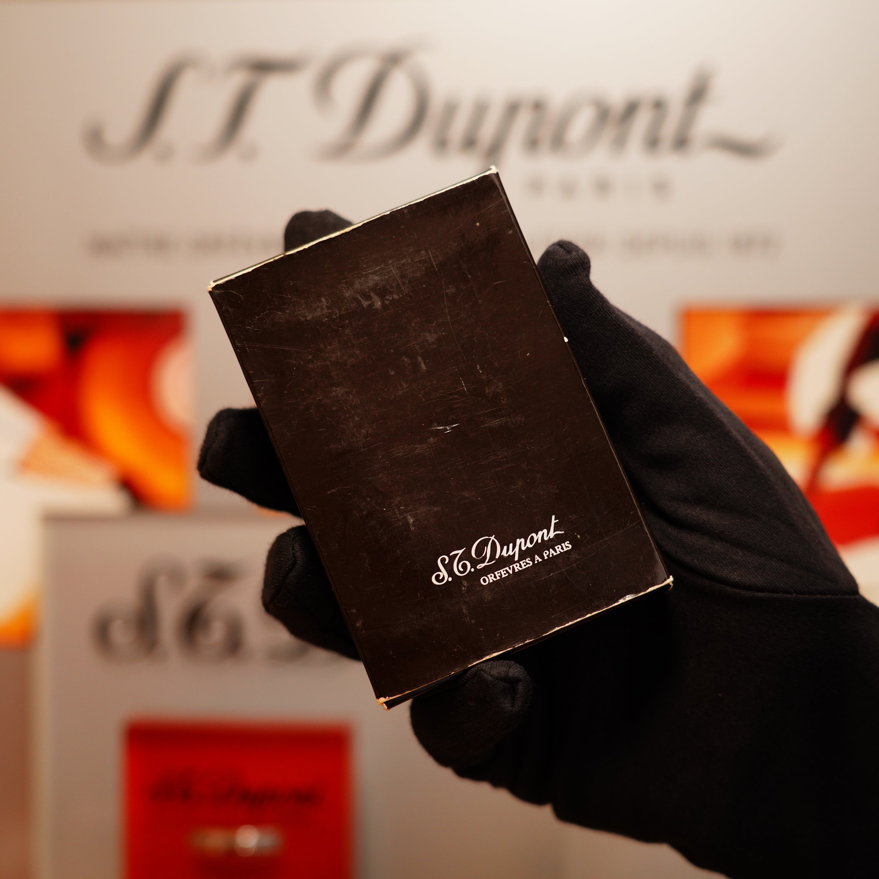 A gloved hand holds a black box with the S.T. Dupont name in front of a blurred background displaying the Vintage 1978 St Dupont 24k Gold Lignes Verticales Pointes Diamants Large Ligne 2 Lighter.
