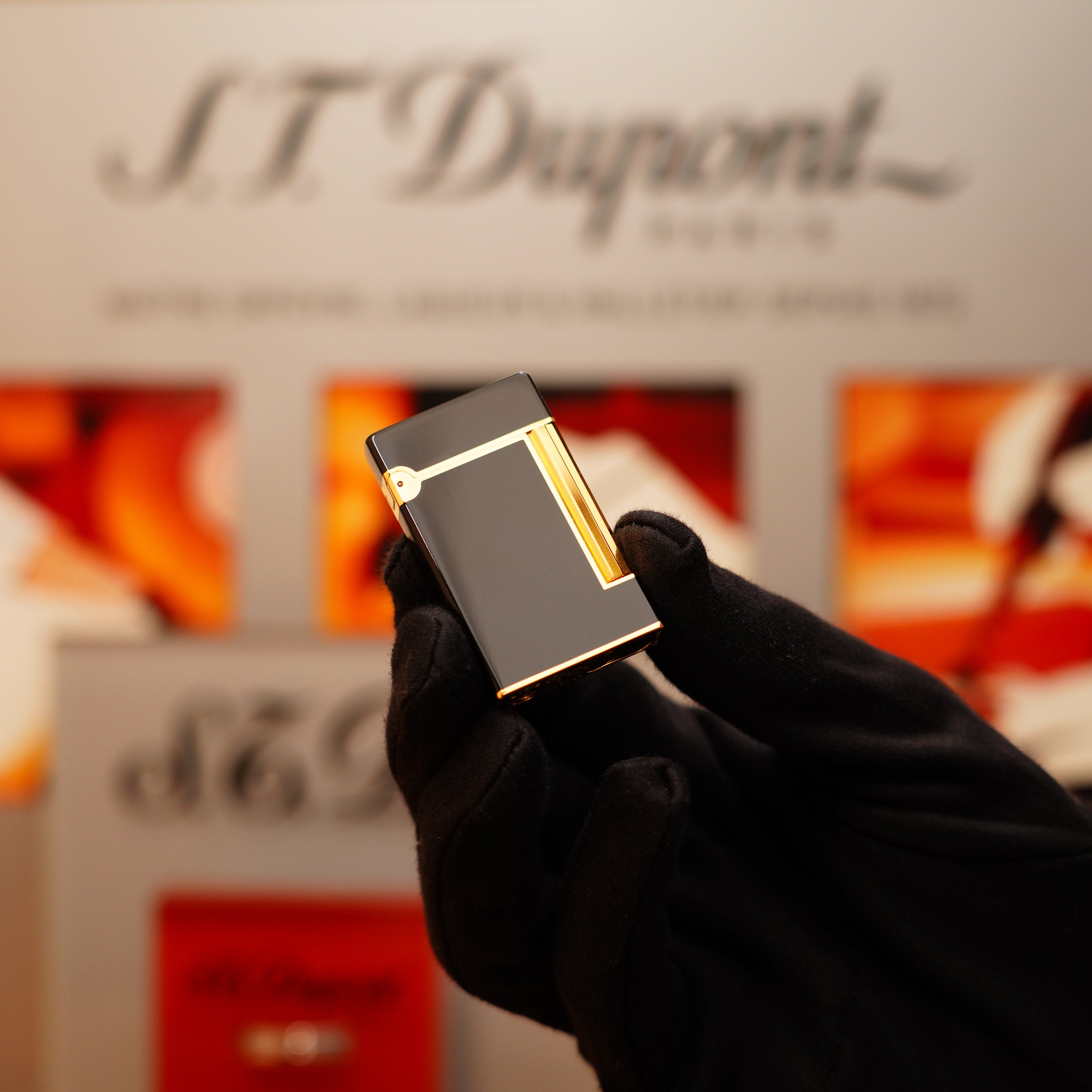 A person in black gloves holds a Vintage 1990 S.T. Dupont Overall Black Lacquer Early 24k Gold Ligne D Lighter indoors, with a blurred logo in the background.