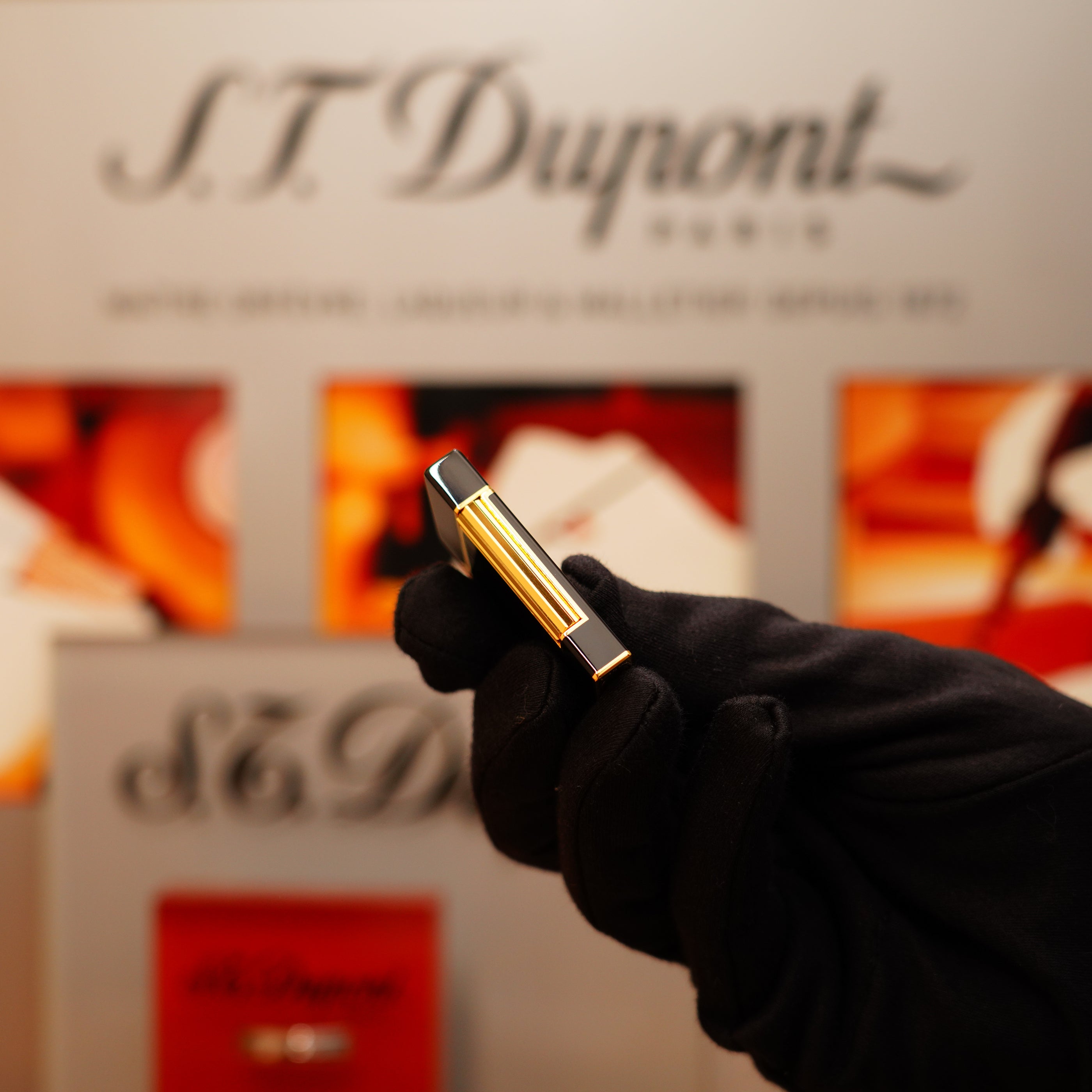 A gloved hand holds a Vintage 1990 St Dupont Overall Black Lacquer Early 24k Gold Ligne D Lighter before a blurred S.T. Dupont display.