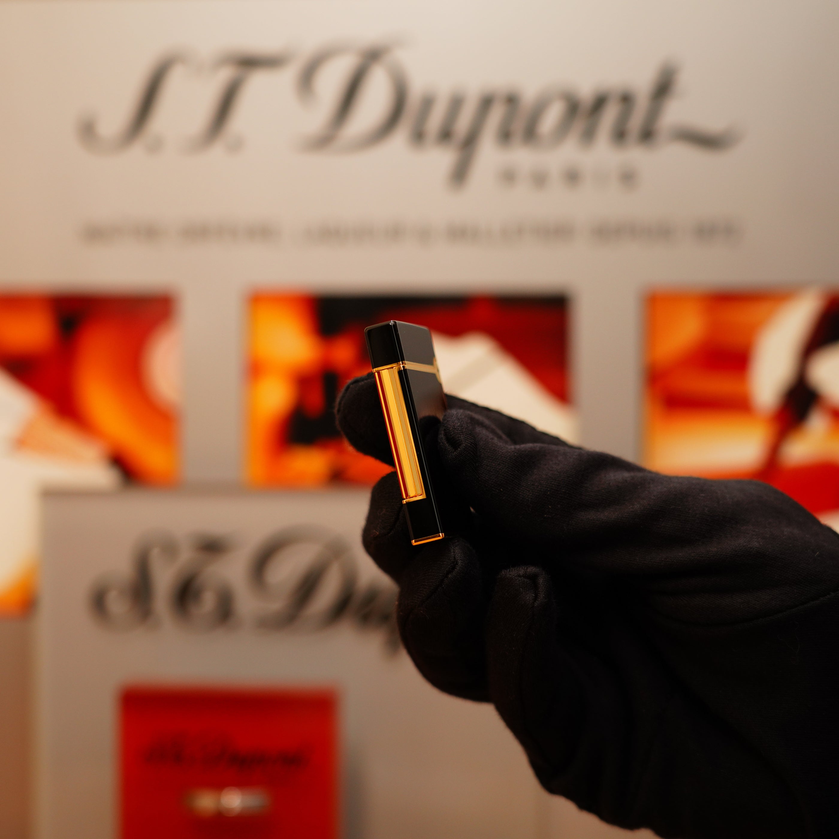 A gloved hand holds a Vintage 1990 S.T. Dupont Overall Black Lacquer Early 24k Gold Ligne D Lighter in front of a blurred S.T. Dupont display.