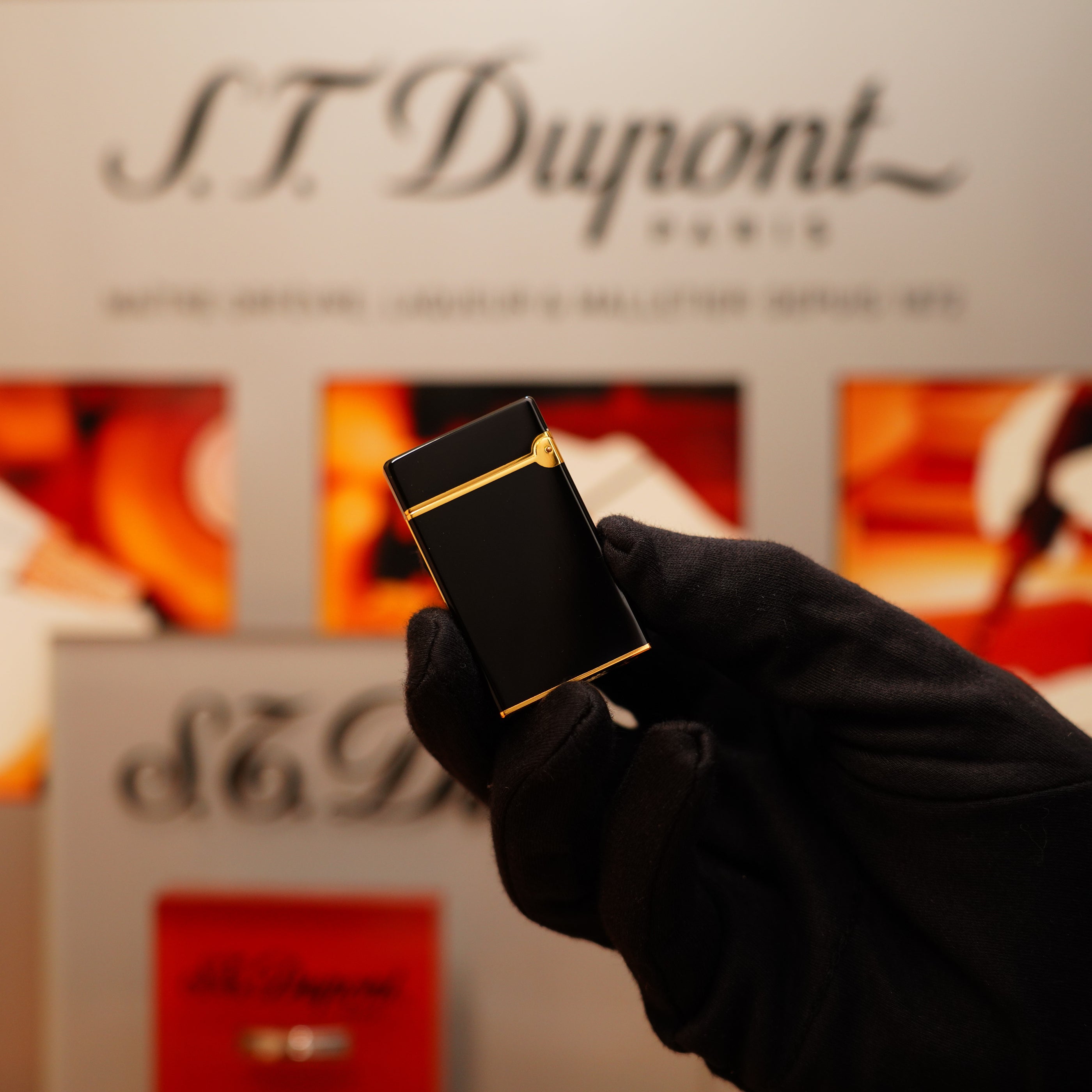 A gloved hand holds a Vintage 1990 S.T. Dupont Overall Black Lacquer Early 24k Gold Ligne D lighter in front of a blurred brand sign.