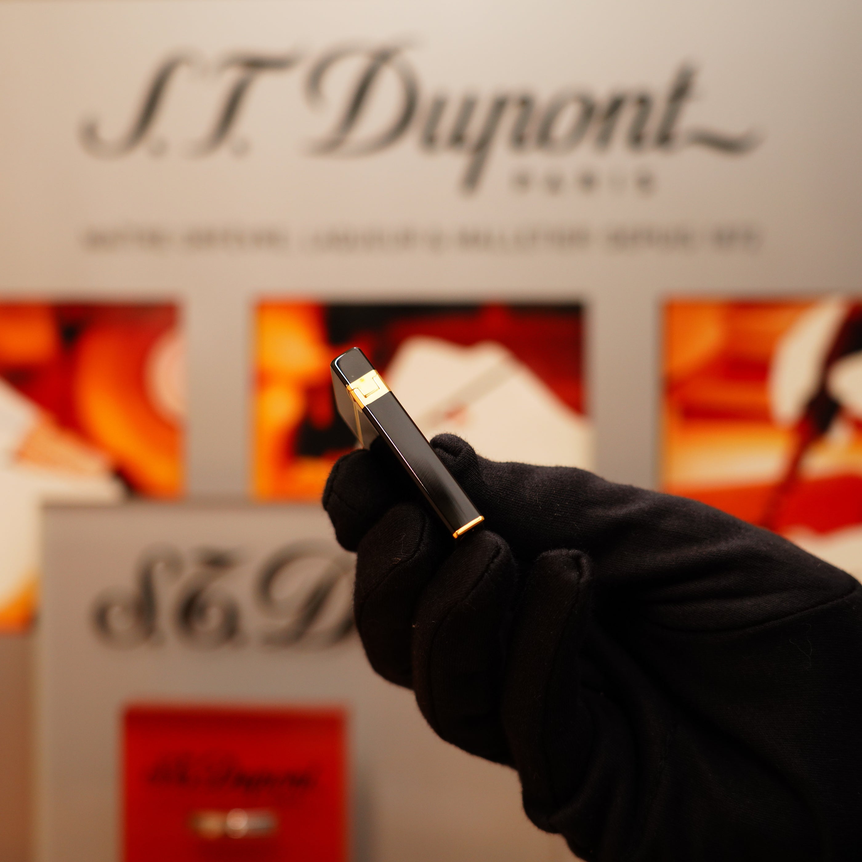 A gloved hand holds a Vintage 1990 S.T. Dupont Overall Black Lacquer Early 24k Gold Ligne D Lighter in front of an S.T. Dupont display.