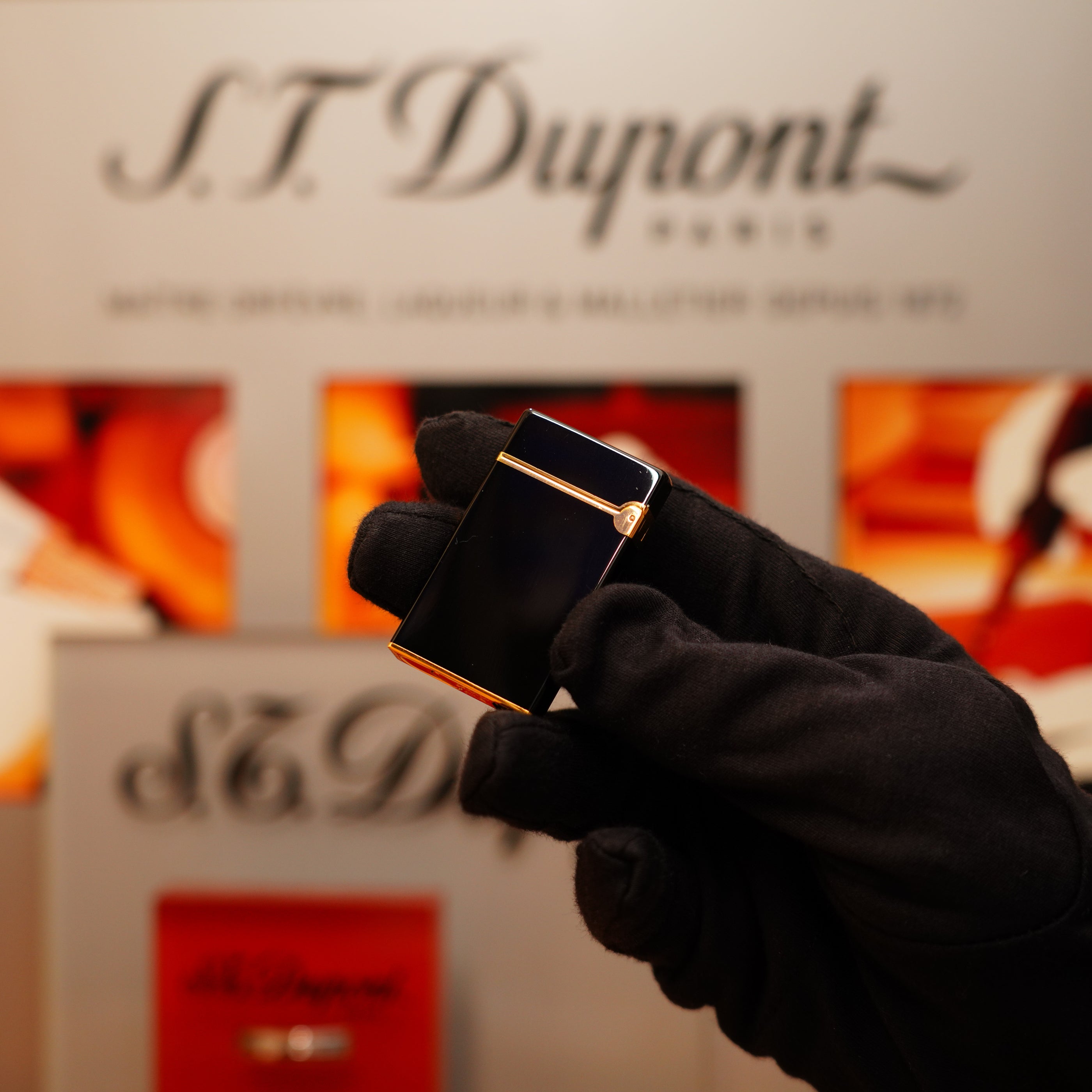 A gloved hand holds a Vintage 1990 S.T. Dupont black lacquer and 24k gold Ligne D lighter in front of an S.T. Dupont display.