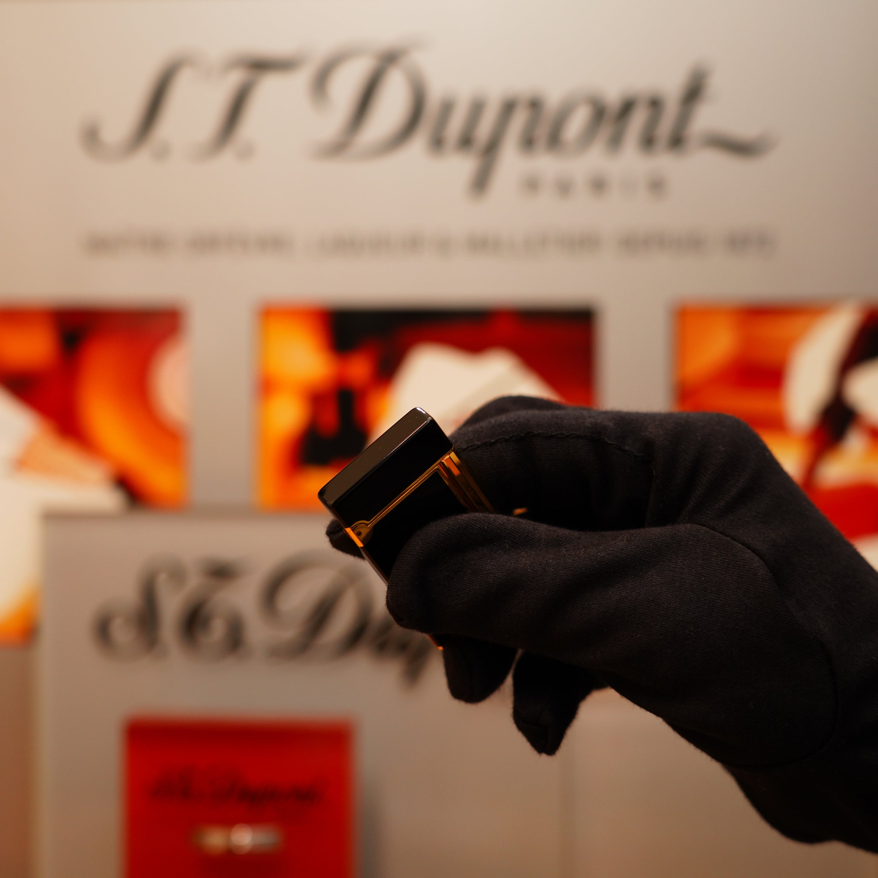 A gloved hand holds a Vintage 1990 St Dupont Overall Black Lacquer Early 24k Gold Ligne D Lighter in front of a blurred S.T. Dupont display.