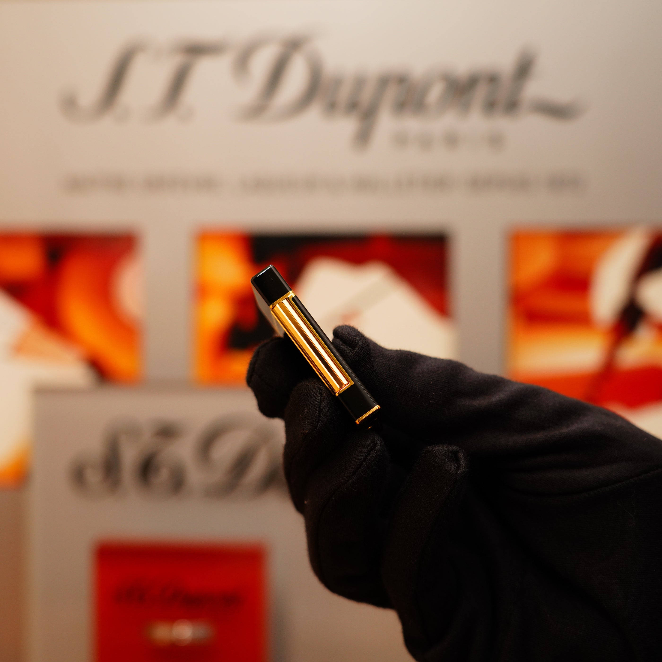 A gloved hand holds a sleek, black and gold vintage 1990 S.T. Dupont Ligne D lighter in front of a blurred S.T. Dupont display.