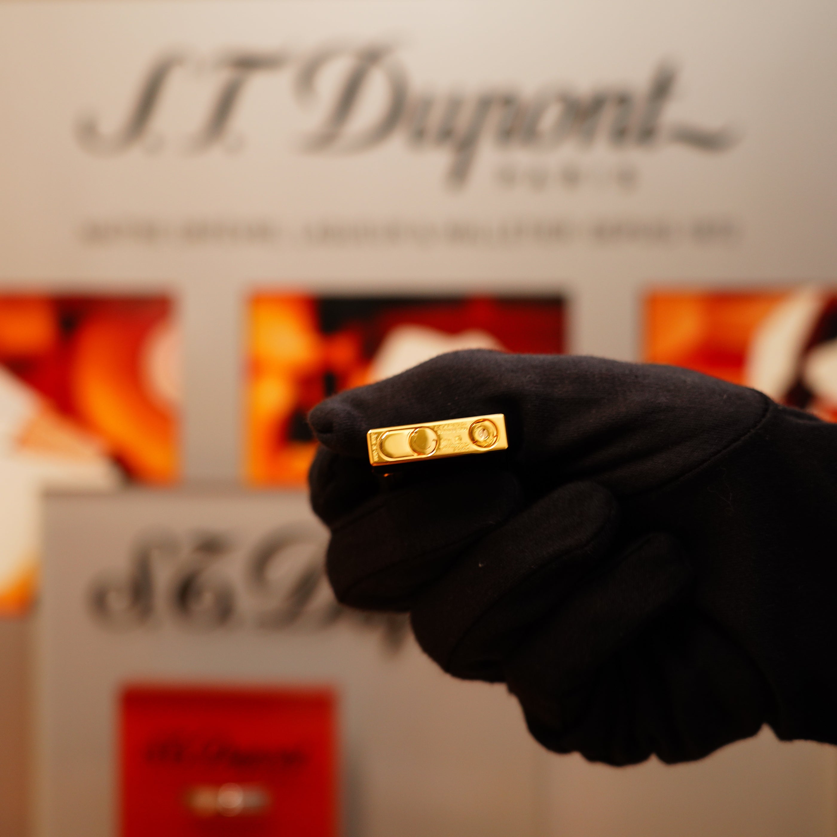 A gloved hand holds a Vintage 1990 S.T. Dupont Overall Black Lacquer Early 24k Gold Ligne D Lighter in front of a blurred backdrop with S.T. Dupont branding.