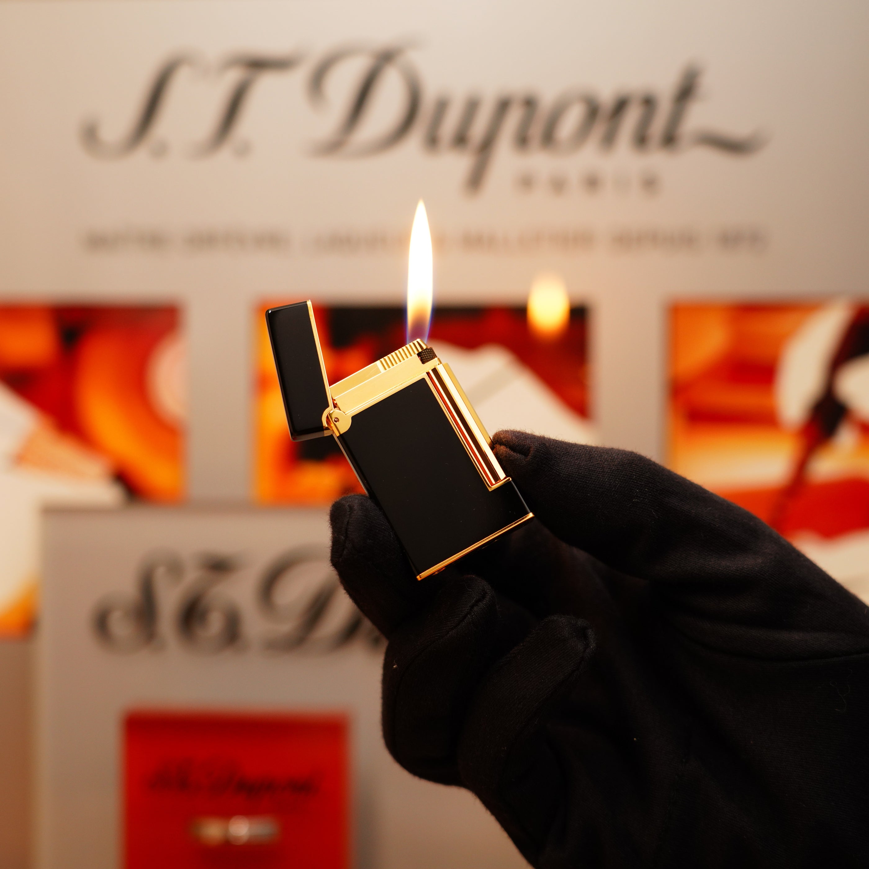 Someone wearing a black glove holds a lit Vintage 1990 S.T. Dupont Overall Black Lacquer Early 24k Gold Ligne D Lighter against a background with blurred branding and text.
