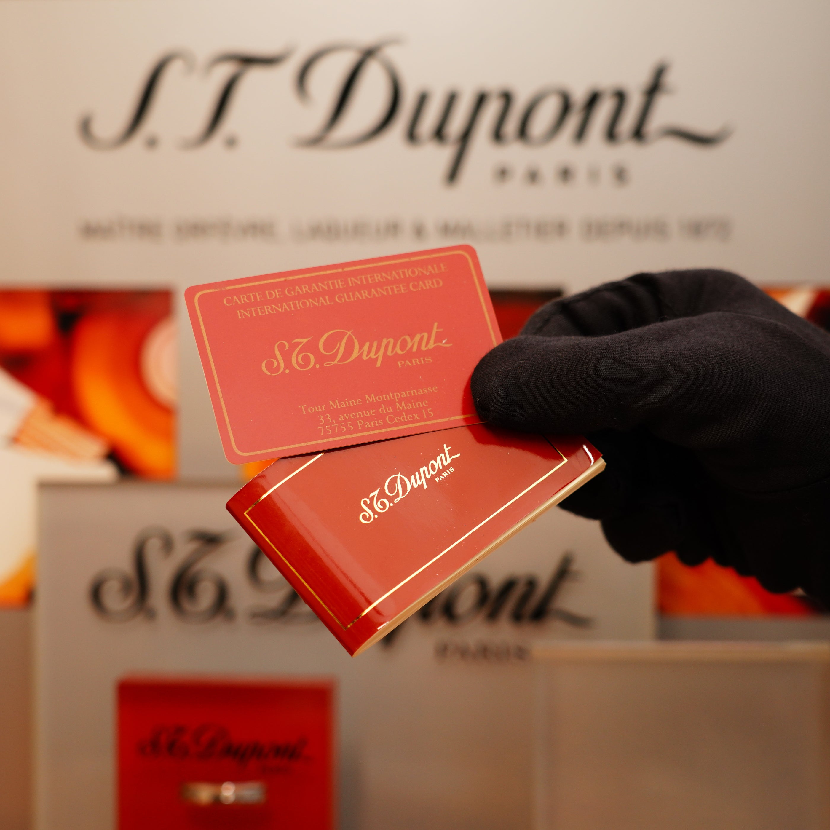 A gloved hand holds a guarantee card from S.T. Dupont above a red box for the Vintage 1990 St Dupont Black Lacquer 24k Gold Ligne D Lighter, set against a branded display.