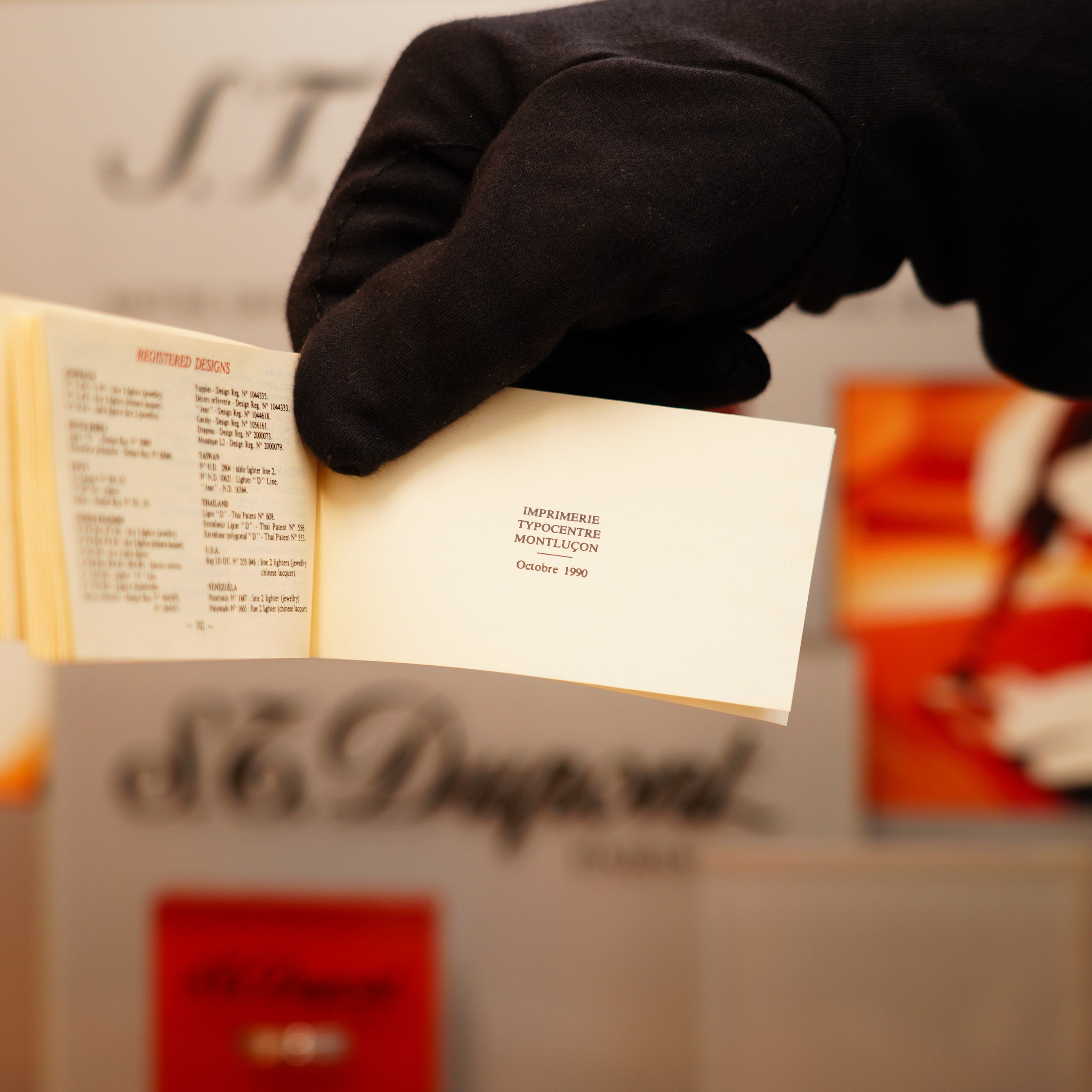 A gloved hand holds a small booklet showcasing "Imprimerie Typographique Montluçon, Octobre 1996," with a blurred background featuring text and orange accents. Nearby, the Vintage 1990 S.T. Dupont Overall Black Lacquer Early 24k Gold Ligne D Lighter stands out in its elegance.