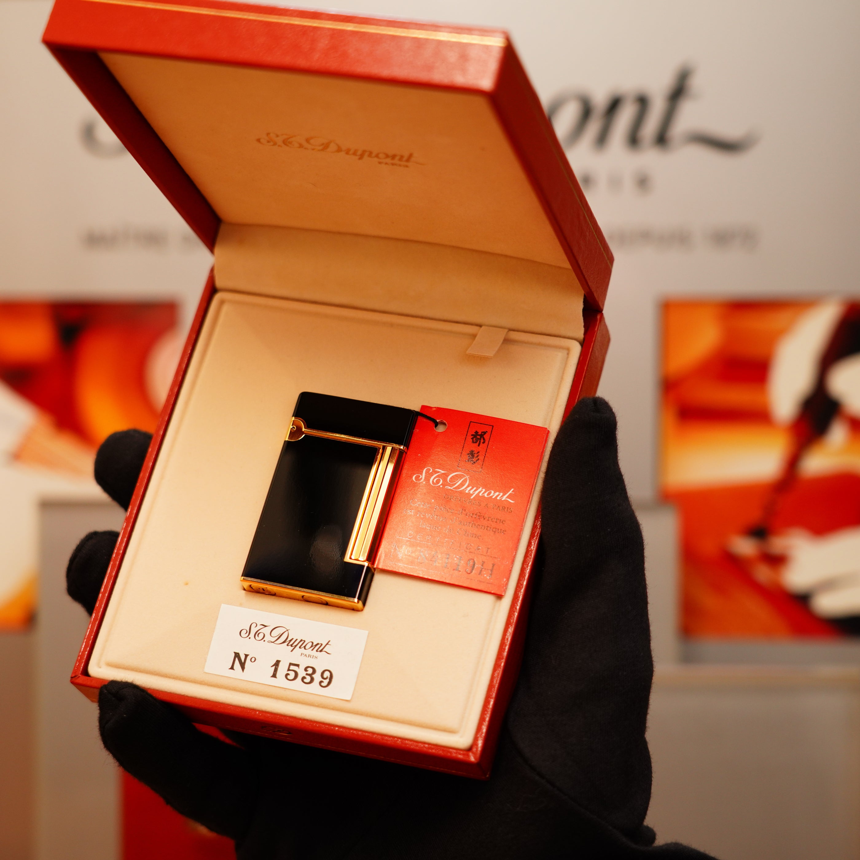 A gloved hand holds a Vintage 1990 S.T. Dupont Overall Black Lacquer Early 24k Gold Ligne D Lighter in an open red box with a label marked "No 1539.
