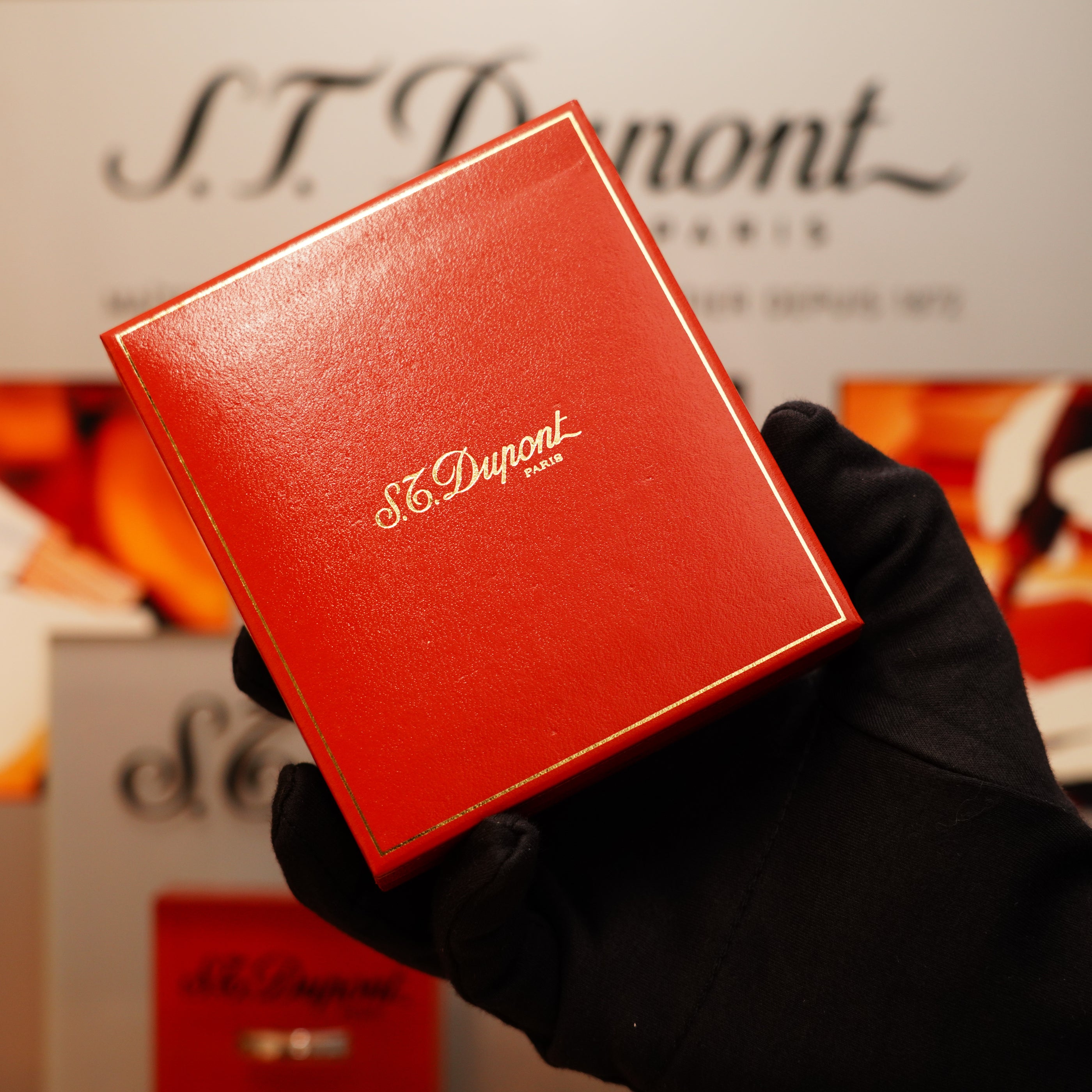A black-gloved hand holds a red S.T. Dupont box with gold script branding, featuring the Vintage 1990 St Dupont Overall Black Lacquer Early 24k Gold Ligne D Lighter, against a blurred backdrop with similar logos.
