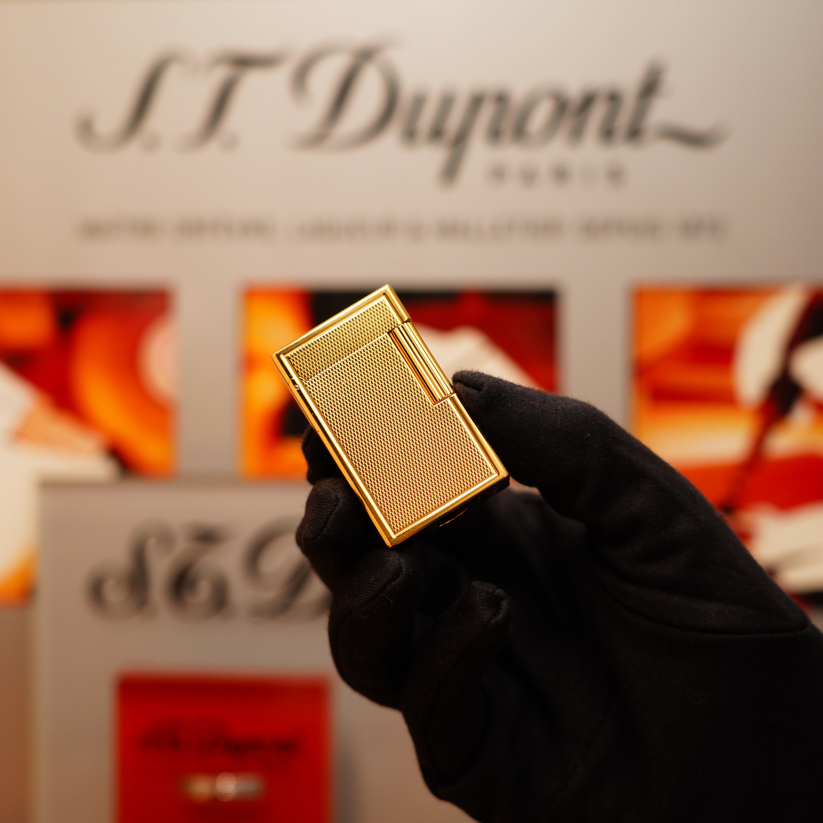 A gloved hand holds a Vintage 1990 S.T. Dupont 24k Large Grain d'Org Iconic Ligne 1 Heavy Finish Lighter against a blurred background displaying the brand's name and red product images.