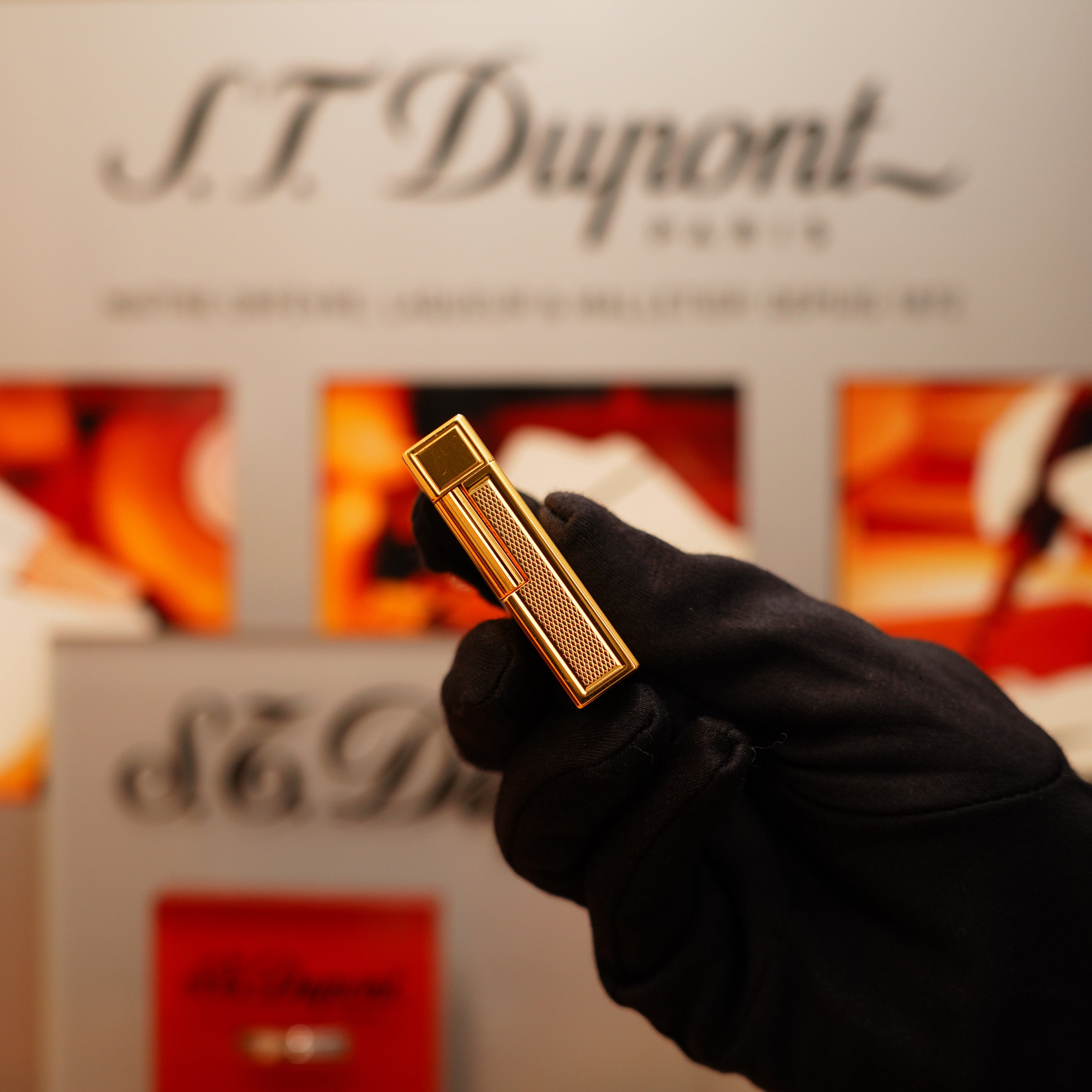 A hand in a black glove holds a Vintage 1990 S.T. Dupont 24k Gold Large Grain d'Org Ligne 1 Heavy Finish Lighter, set against a blurred S.T. Dupont display.