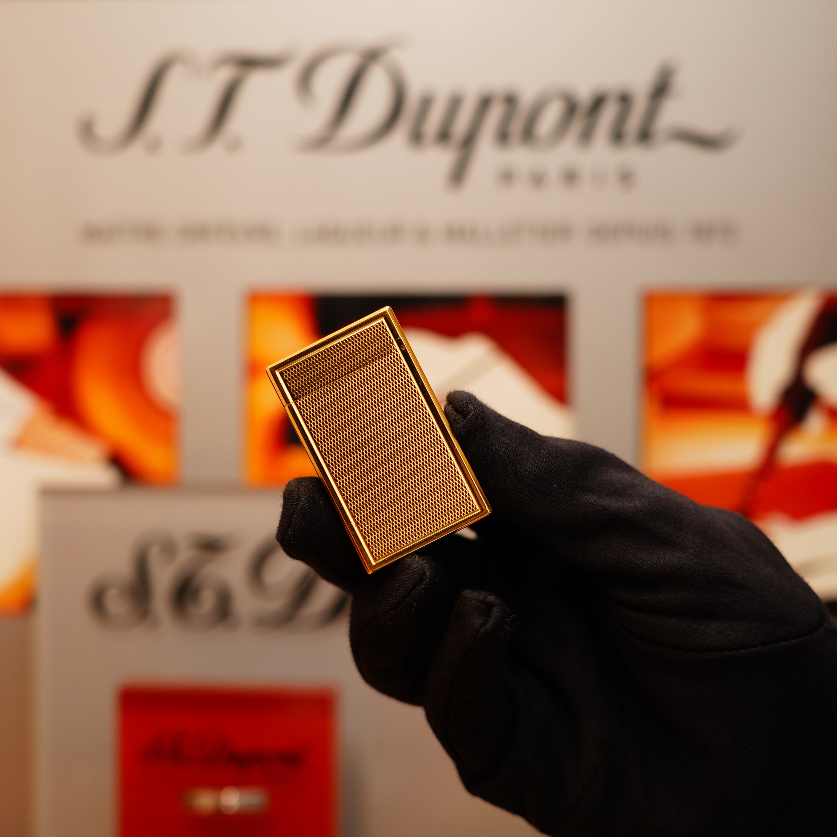 A gloved hand holds a gold-colored Vintage 1990 S.T. Dupont 24k Large Grain d'Org Iconic Ligne 1 Heavy Finish Lighter in front of a blurred S.T. Dupont Paris sign.