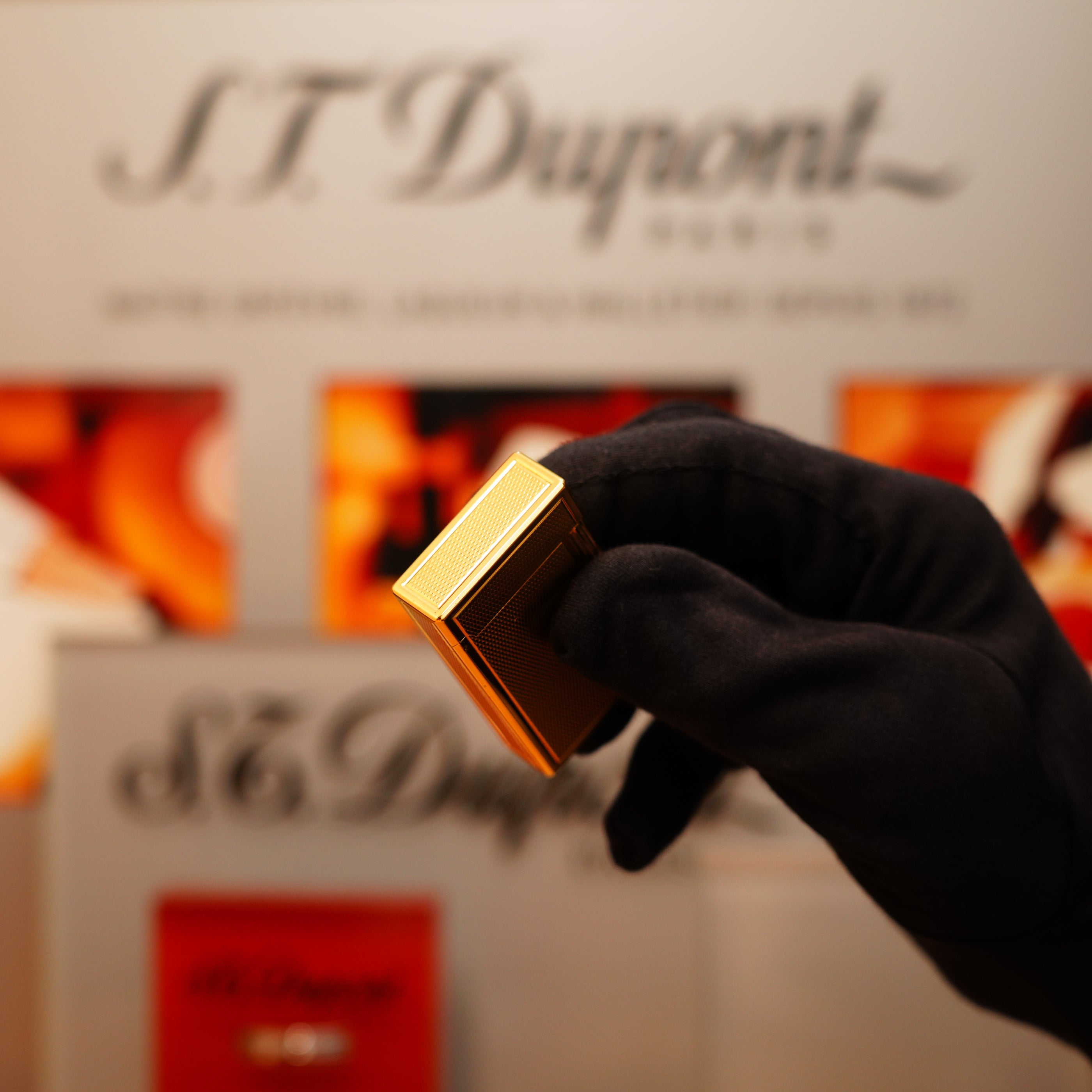 A gloved hand holds a Vintage 1990 S.T. Dupont 24k Large Grain d'Org Iconic Ligne 1 Heavy Finish Lighter against a blurred background featuring the S.T. Dupont logo.